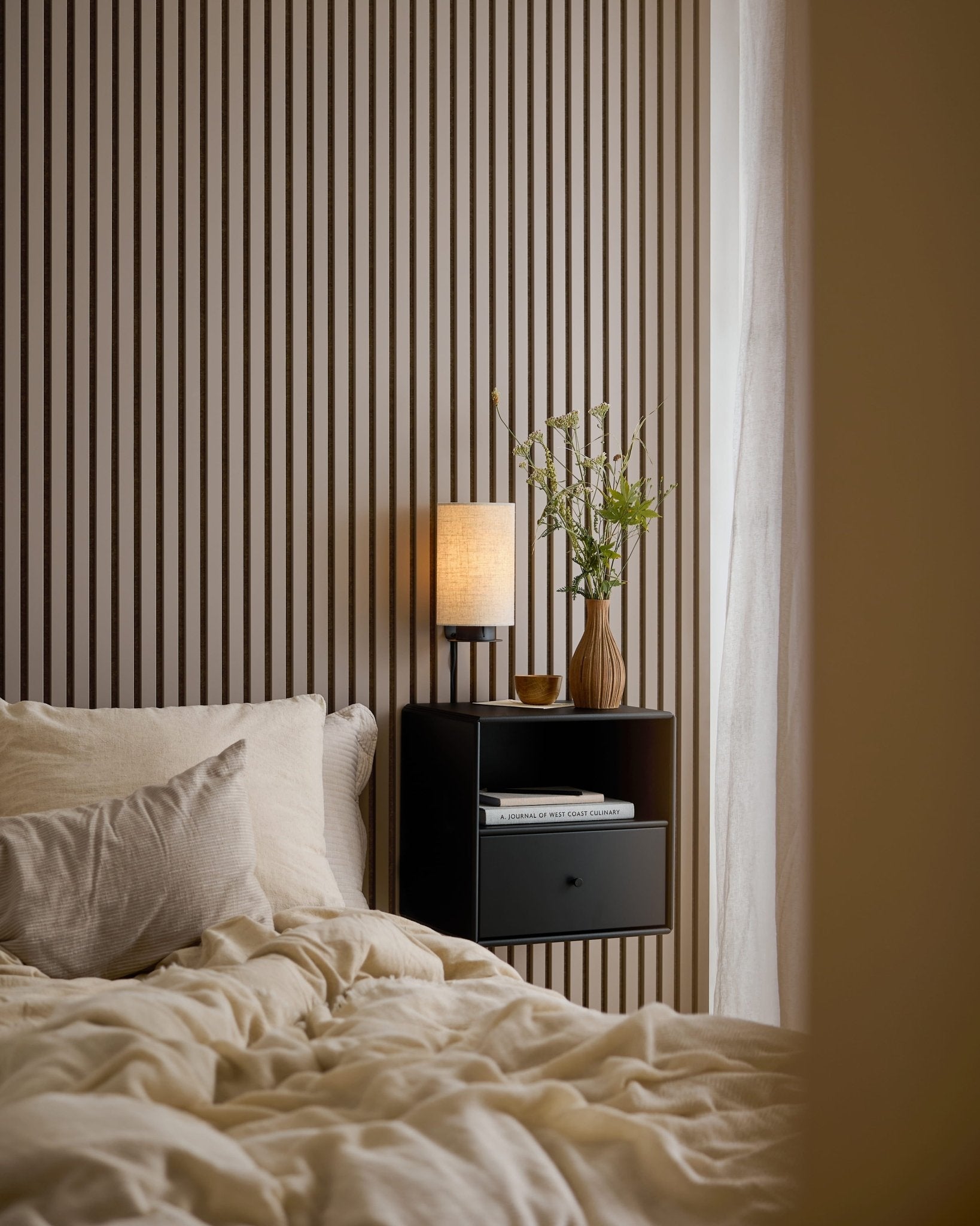 Un lit habillé de linge crème à côté d'une table de chevet noire, devant un mur Akupanel en Dark Cashmere.