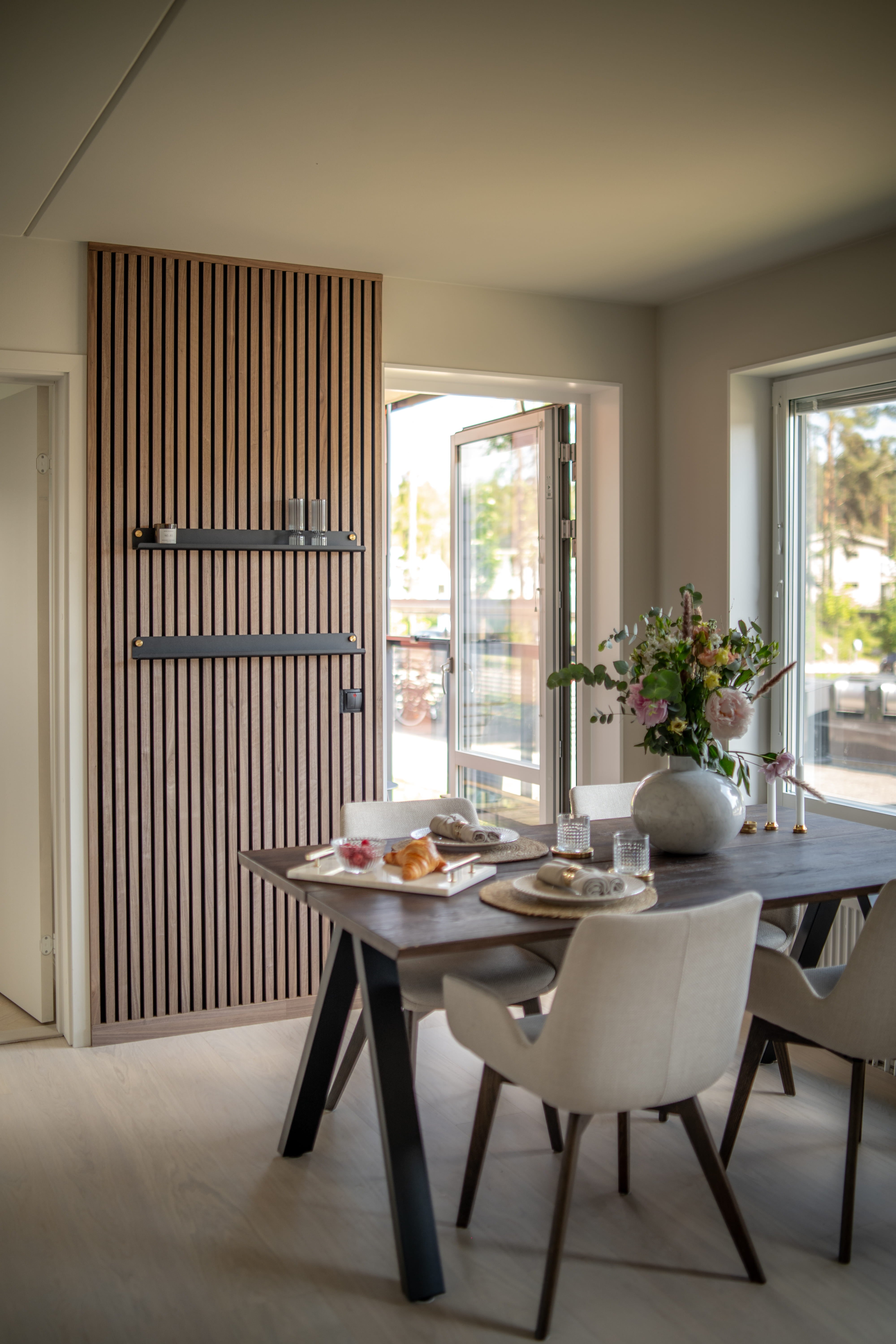 Salle à manger moderne avec un mur de lattes en bois acoustique Akupanel texturé, une table de petit-déjeuner sombre, des chaises minimalistes beiges et un vase de fleurs près de grandes fenêtres ouvertes.