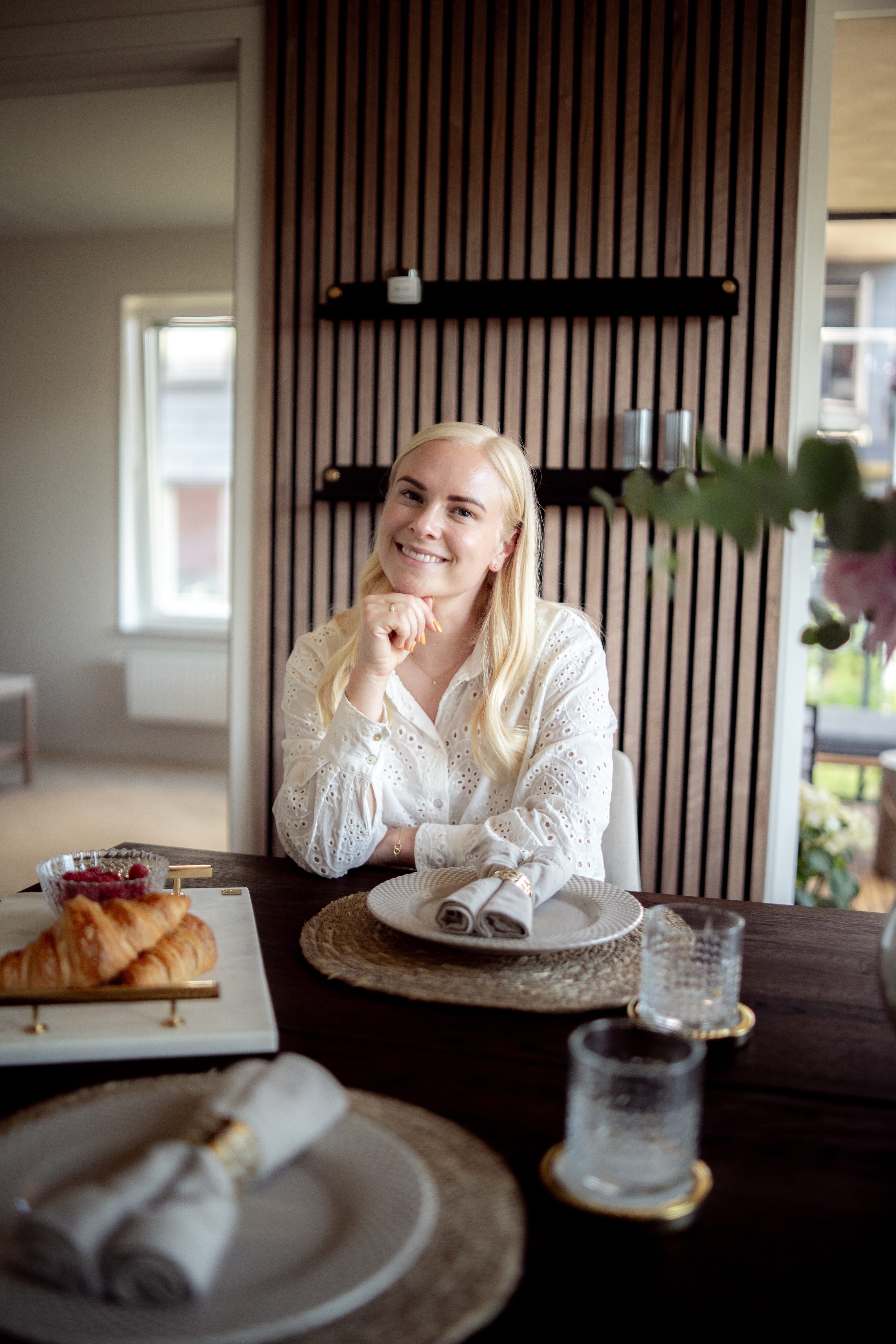 Une femme aux longs cheveux blonds est assise à une table préparée pour le petit-déjeuner, avec des croissants et des verres, à côté dun panneau acoustique nordique accroché au mur.