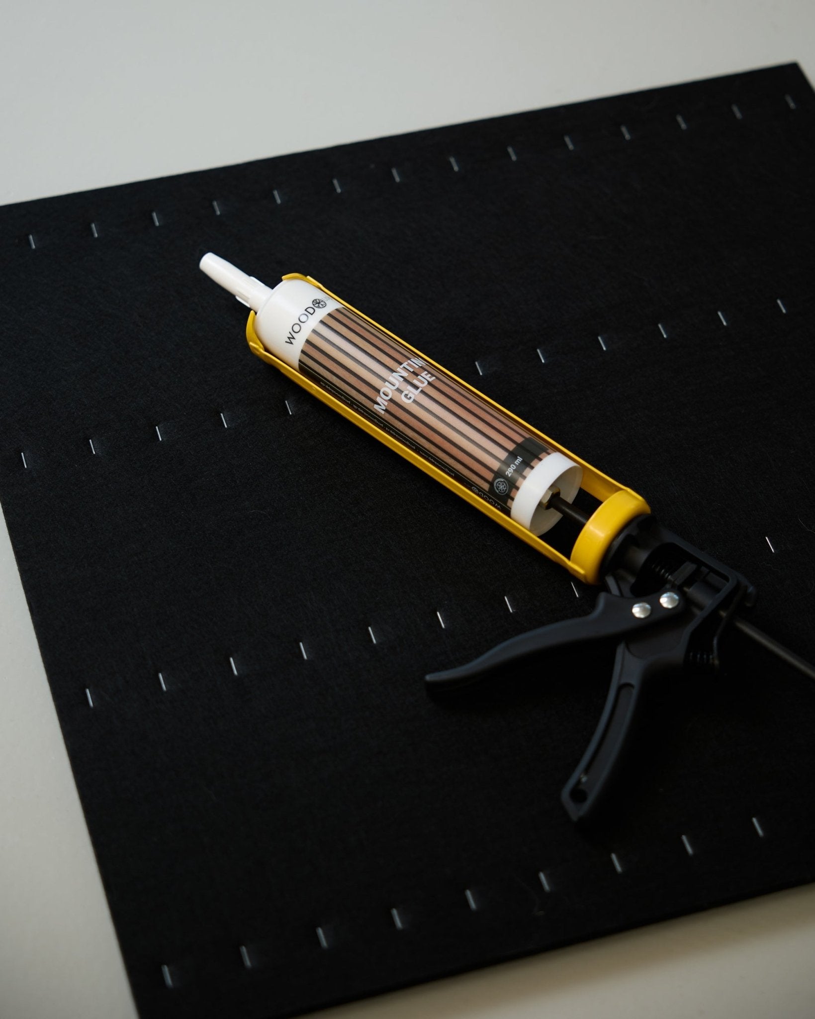 Un pistolet à colle WoodUpp jaune et black (290 ml) repose sur un tapis black avec une grille d’agrafes blanches, utilisé pour installer des panneaux acoustiques.