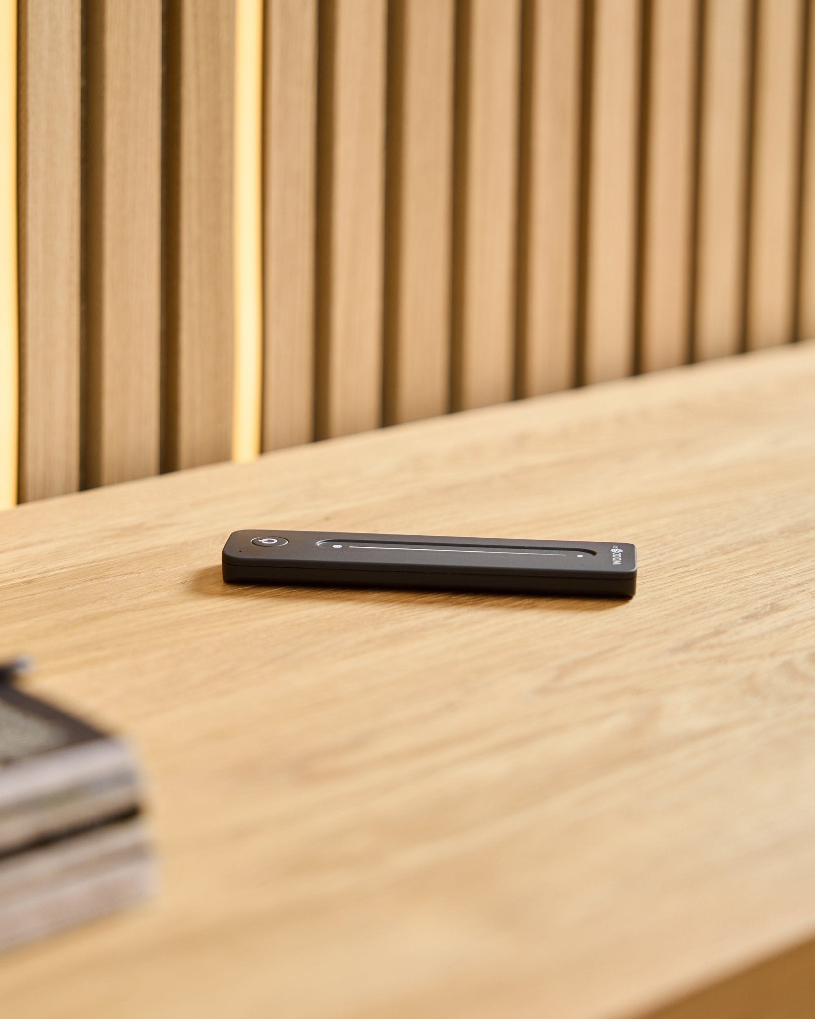 Une télécommande LED WoodUpp black Lumi posée sur une surface en bois clair, avec en arrière-plan des panneaux acoustiques en bois à lattes.