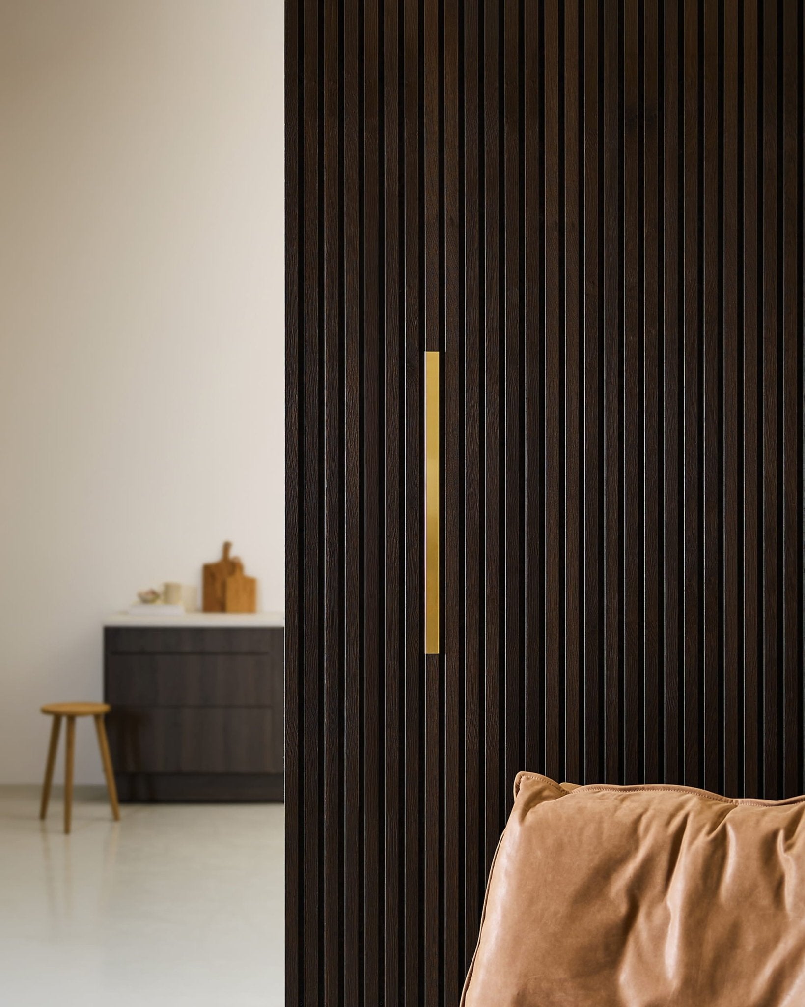 Pièce minimaliste mettant en valeur un panneau WoodUpp Akupanel Liva | Revêtement / Laiton, 60 cm, à côté dun fauteuil en cuir, dun tabouret et dun meuble sombre, dans un décor neutre.