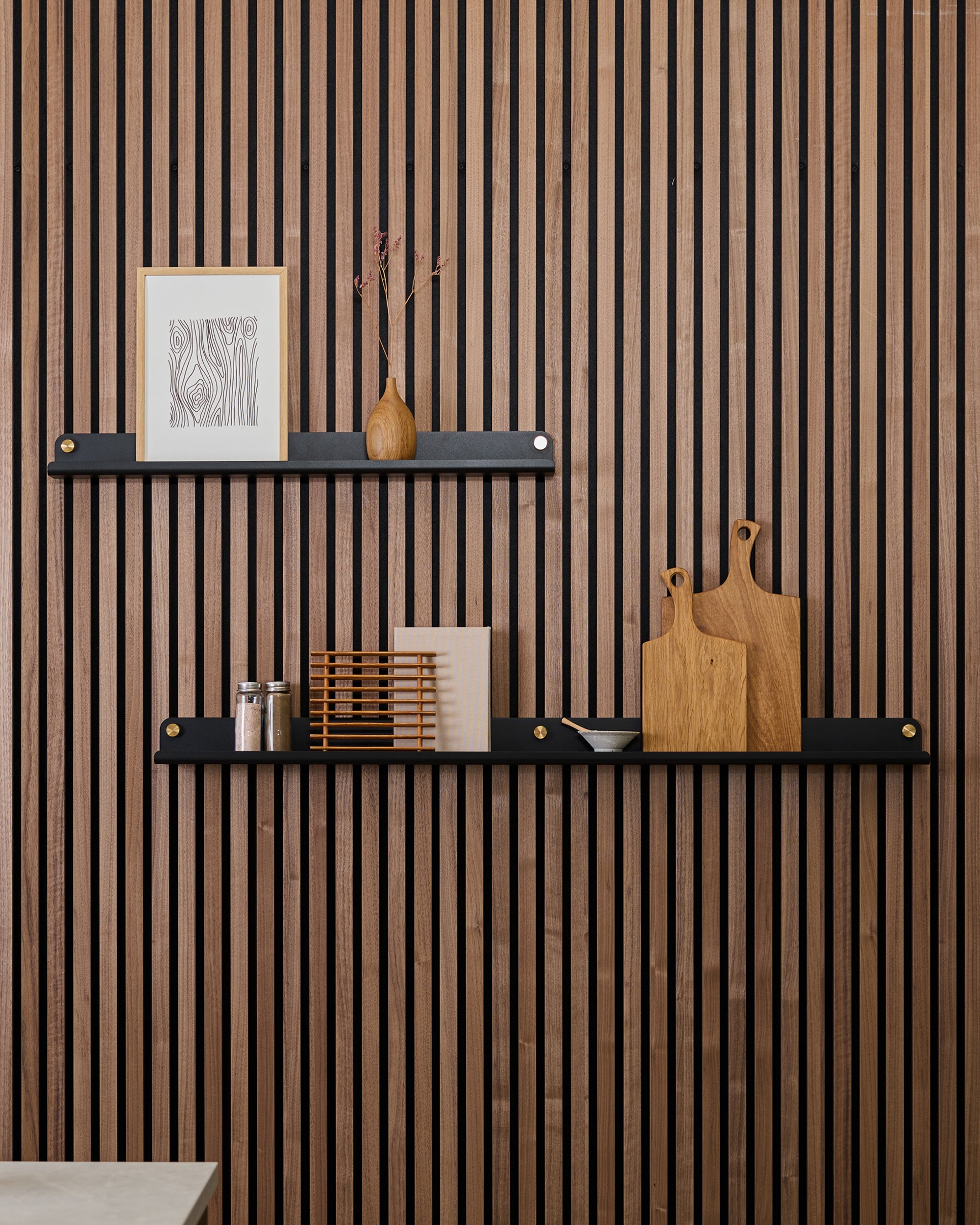 Two black wall shelves mounted on a vertical textured acoustic wood slat panel, displaying art, a vase, kitchen jars, spice rack, and wooden cutting boards.