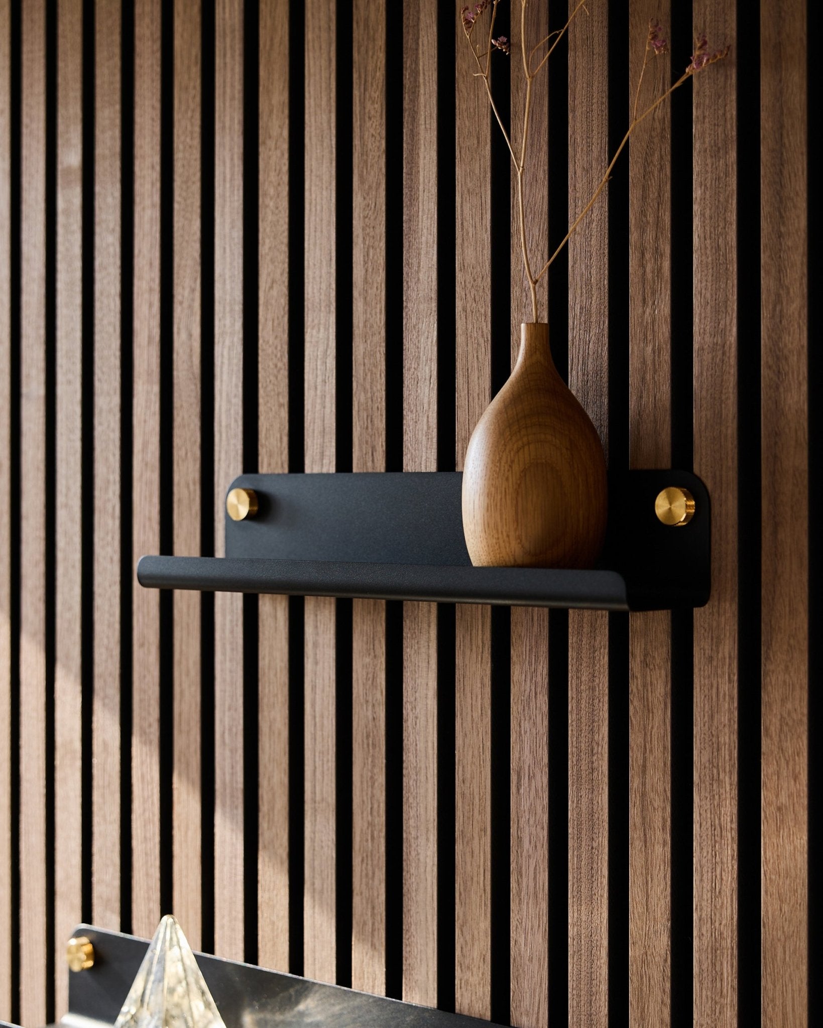 Un vase en bois avec des branches séchées sur une étagère étroite WoodUpp black Norr (35,9 cm), devant un panneau acoustique à lattes de style nordique vertical.