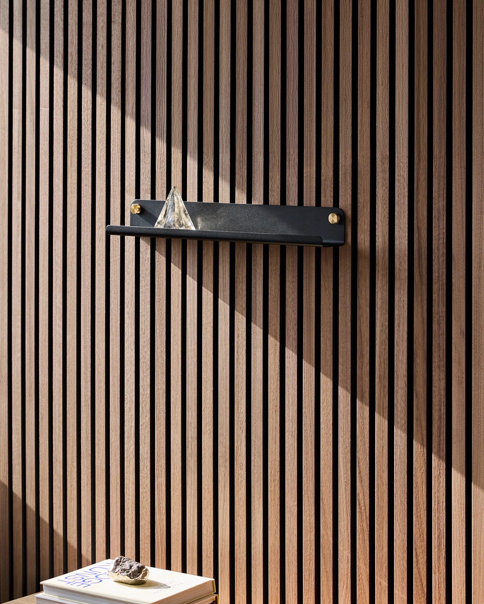 Une étagère Norr Narrow noire de la marque WoodUpp, posée sur un panneau acoustique en bois Akupanel, accueille une pyramide en verre ; la lumière du soleil illumine les livres situés en dessous, surmontés dun caillou.