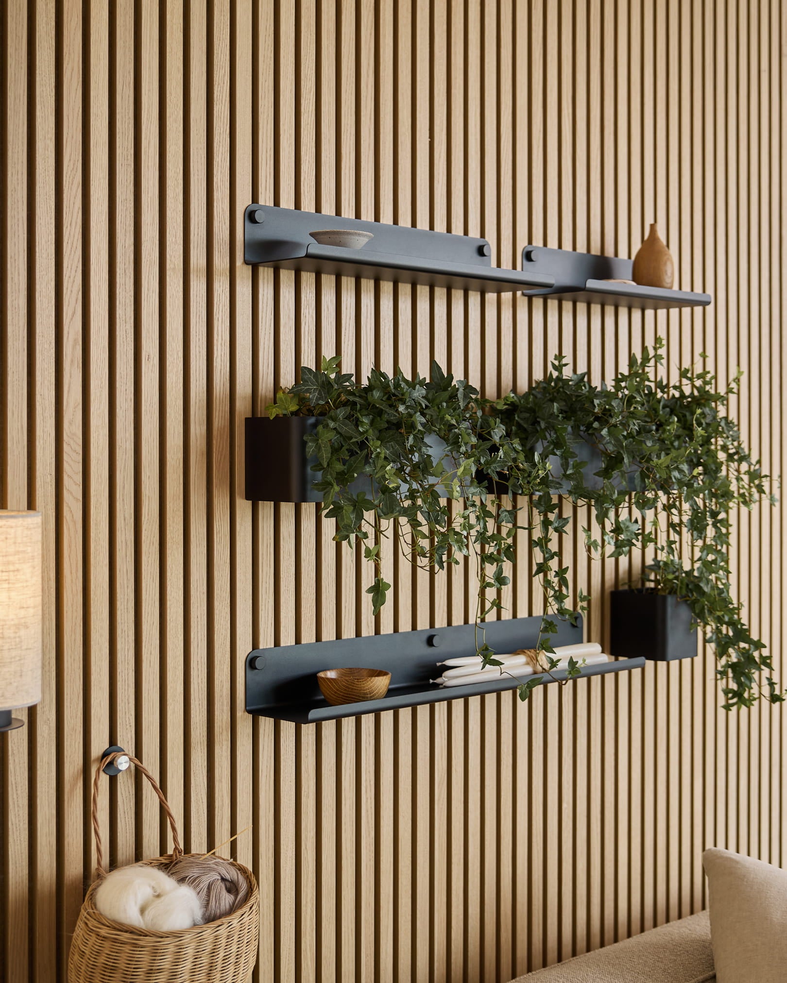 Trois étagères black Norr | Plant Box de WoodUpp, avec des objets déco et une plante, sont fixées au-dessus d’un panier en osier rempli de pelotes, sur fond de panneau acoustique mural à lattes vert.