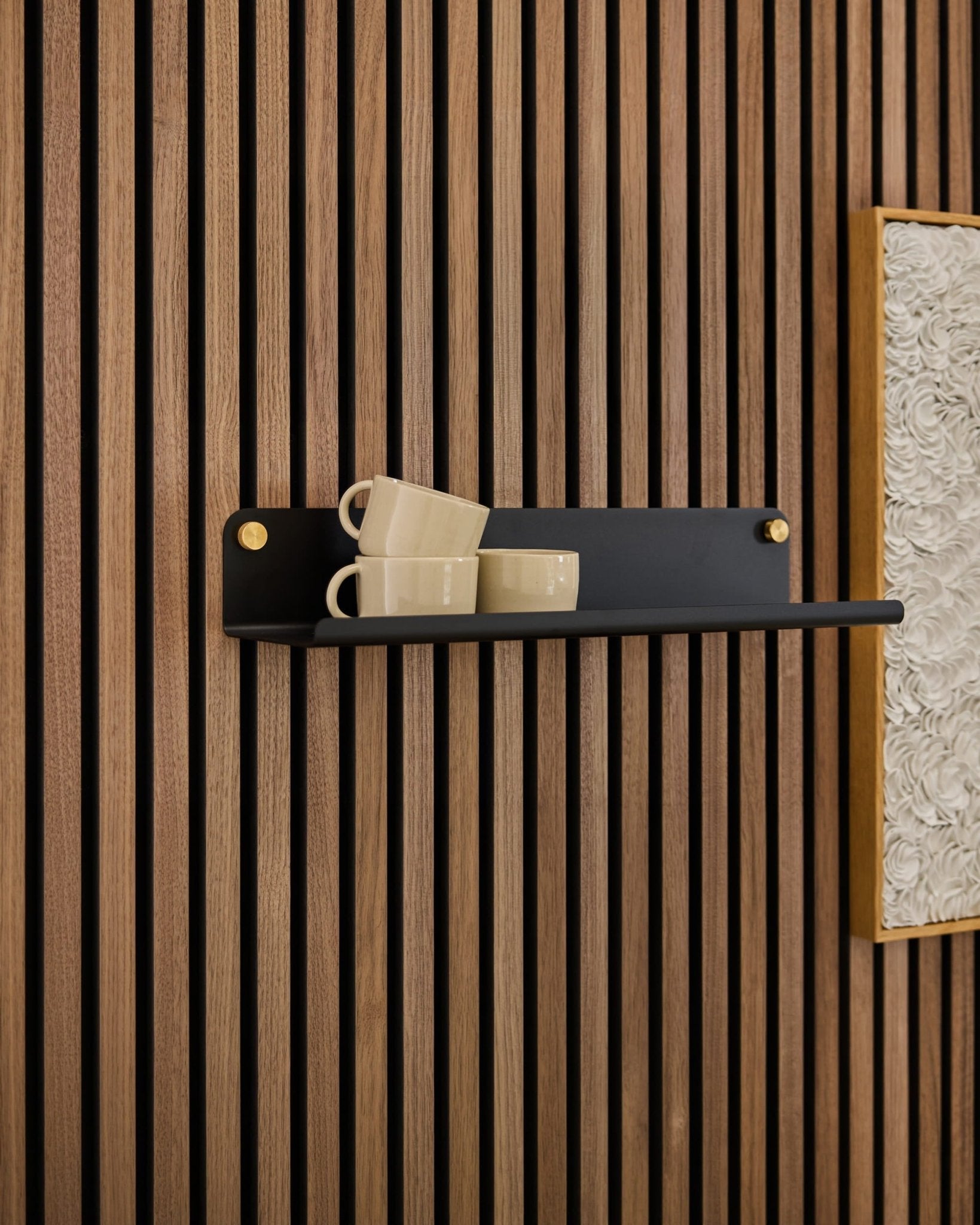 Une étagère large Norr noire de la marque WoodUpp accueille trois tasses beiges, posées sur un panneau acoustique Akupanel, à côté dune œuvre dart blanche texturée encadrée.