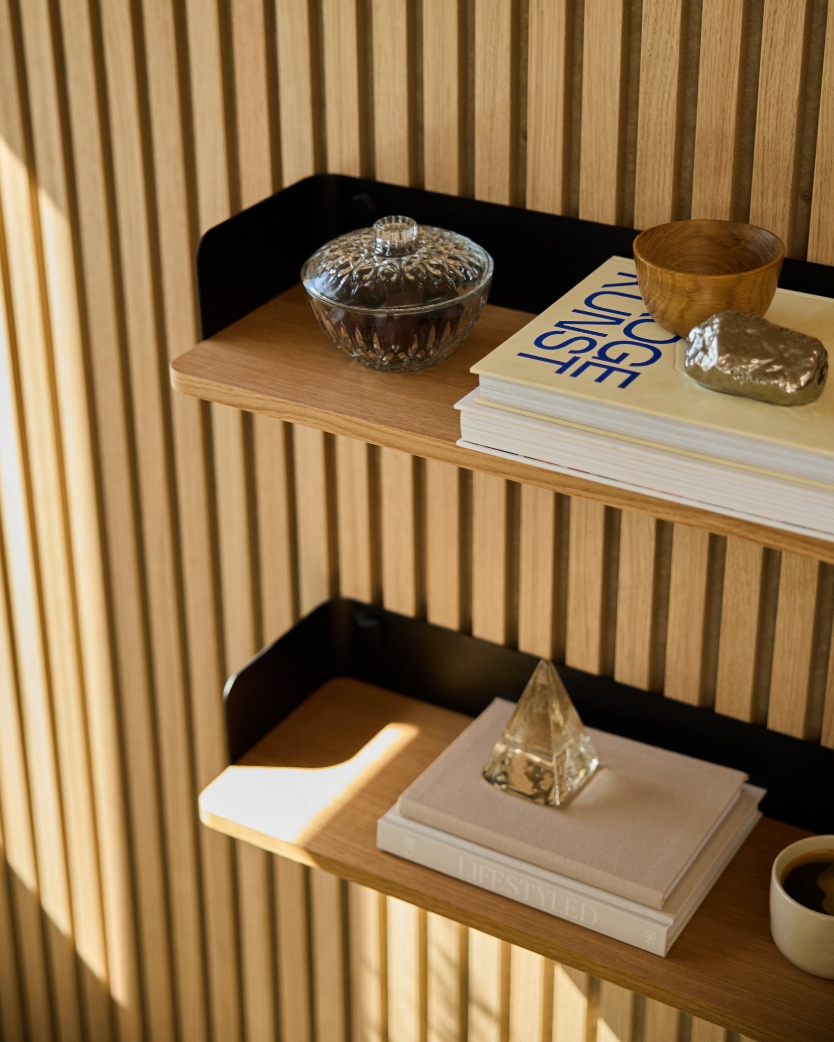 La bibliothèque WoodUpp Linn de Natural Oak (55,9 cm) accueille un bocal en verre, une tasse, une pyramide dorée et des livres devant des panneaux acoustiques en bois Akupanel, baignés de soleil.