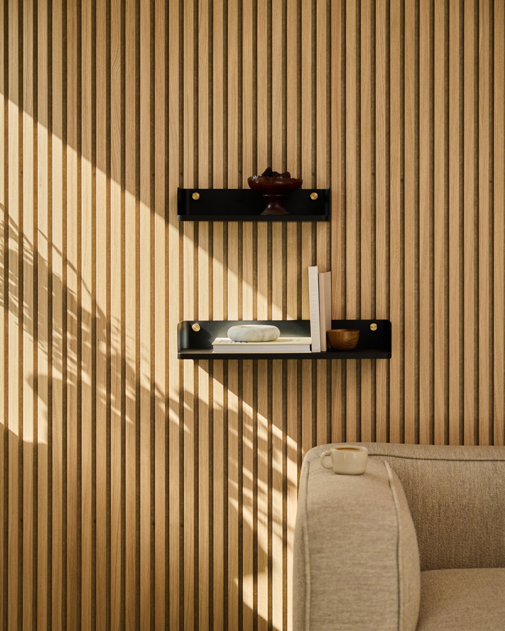 La lumière du soleil éclaire un canapé beige et la bibliothèque Linn de WoodUpp en black oak, 55,9 cm, devant un panneau mural à lattes nordique minimaliste Panneau acoustique.