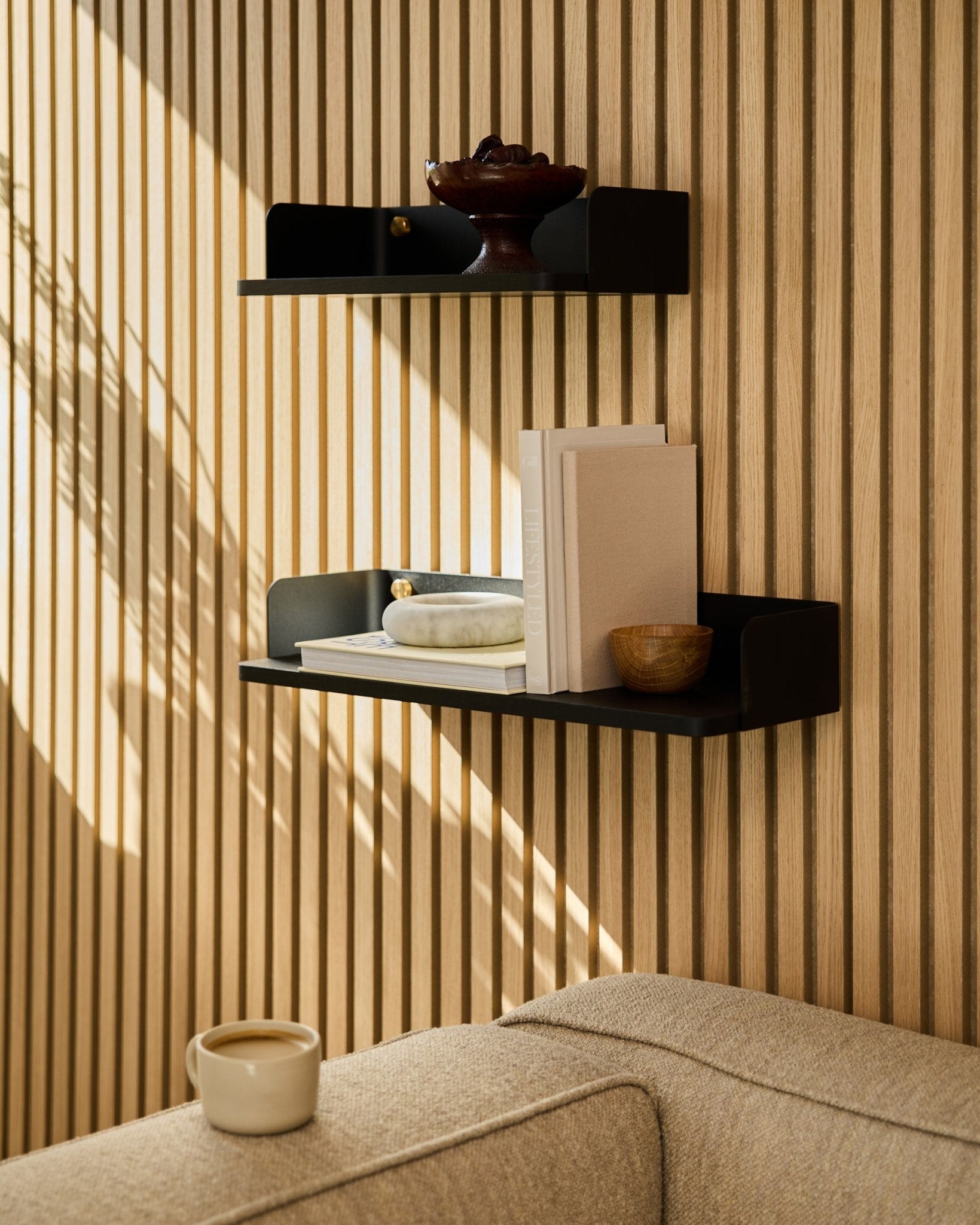 Une bibliothèque WoodUpp black oak (55,9 cm) présente des livres, des objets déco et un bol sur un mur en panneaux acoustiques en bois ; un canapé beige et une tasse au premier plan.