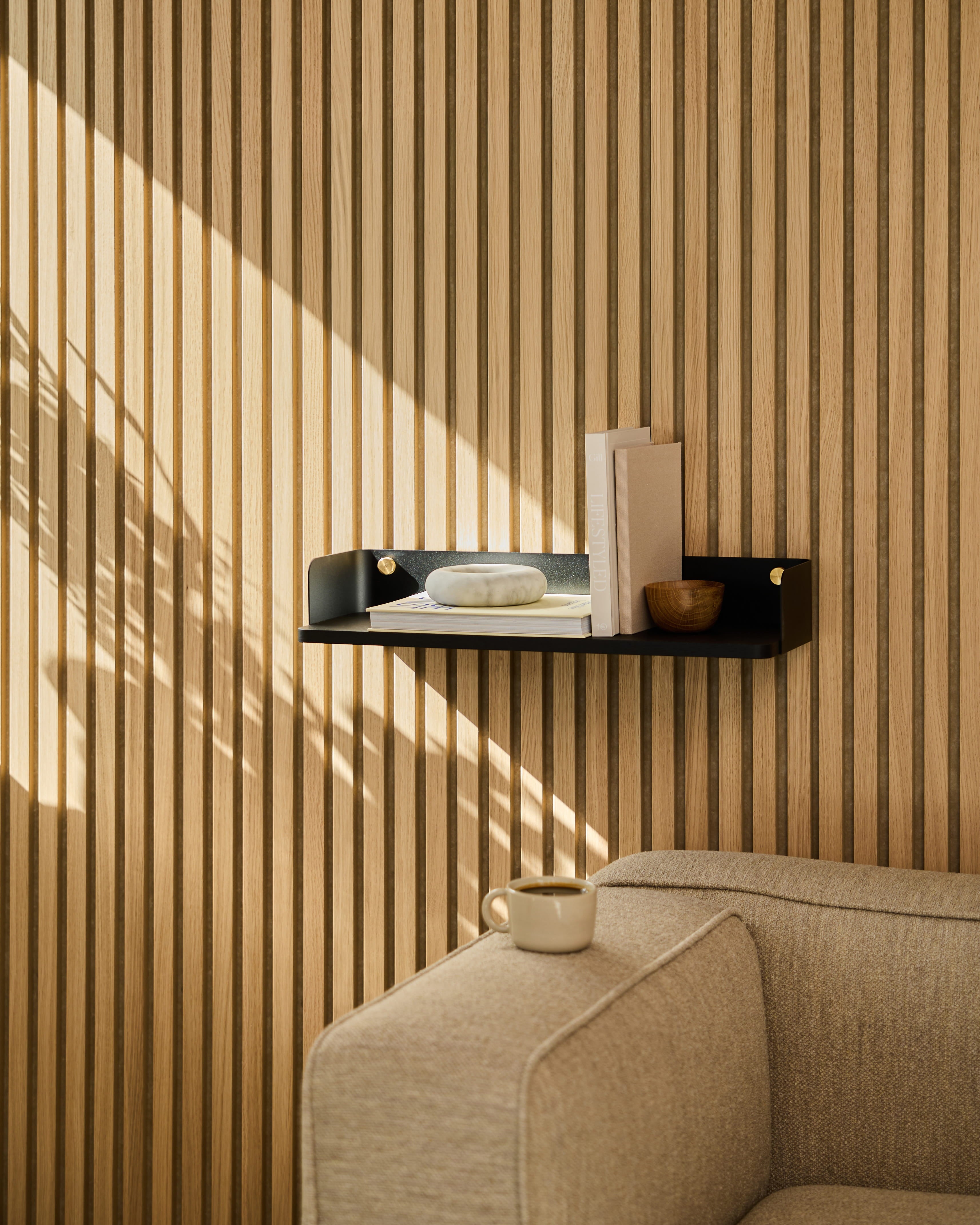 Une étagère A Linn | Bookshelf / Black Oak de WoodUpp met en valeur la décoration au-dessus dun canapé beige. La lumière du soleil projette des ombres sur le mur recouvert de panneaux acoustiques en bois rayés.
