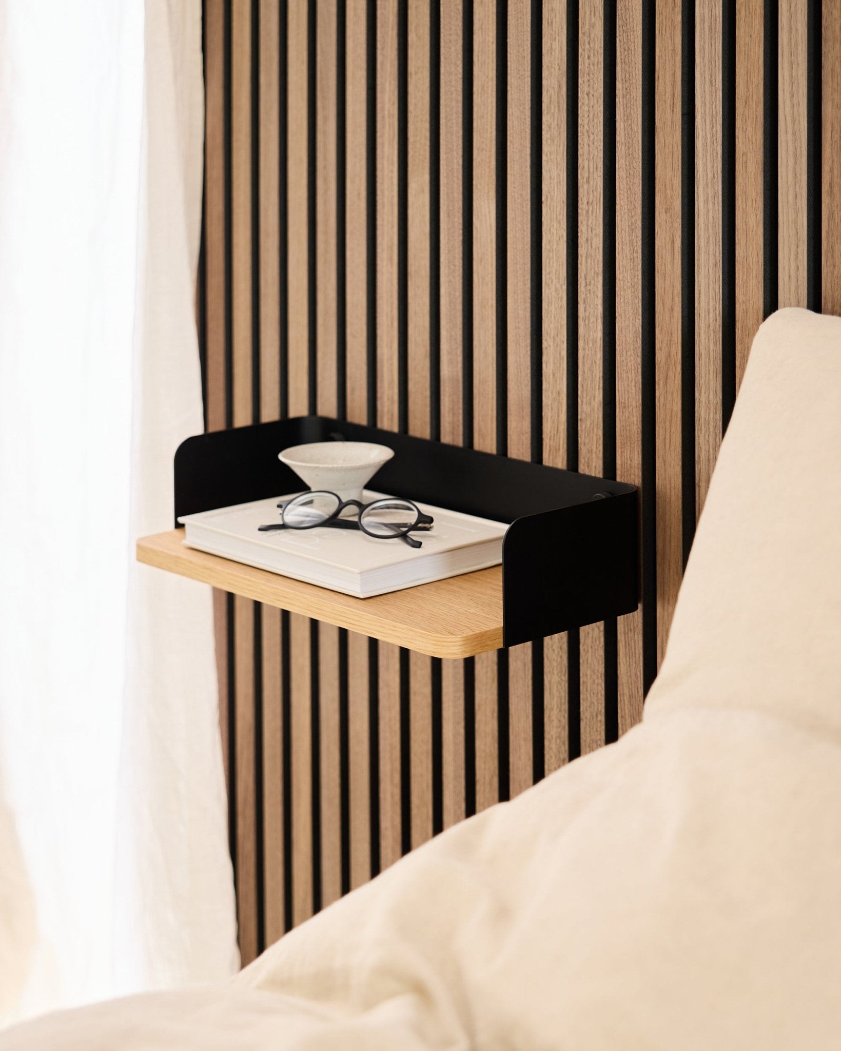 La bibliothèque Linn de WoodUpp en natural oak (39,9 cm), avec un livre, des lunettes et un verre, posée à côté d’un lit devant un mur en panneaux acoustiques en bois Akupanel.