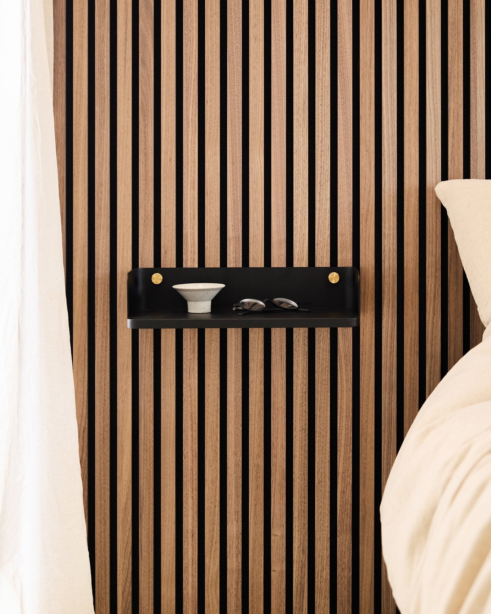 L’étagère Linn en Black Oak de WoodUpp est montée sur un panneau acoustique à lattes au style nordique et accueille un bol et des verres à côté d’un lit avec du linge de lit beige.