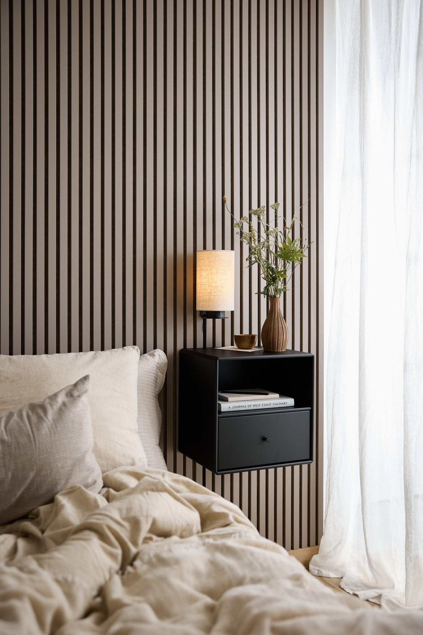 Un lit avec une parure de lit beige, une commode-bibliothèque WoodUpp Arka noire, une lampe et un vase près dune fenêtre ornée de rideaux blancs vaporeux.