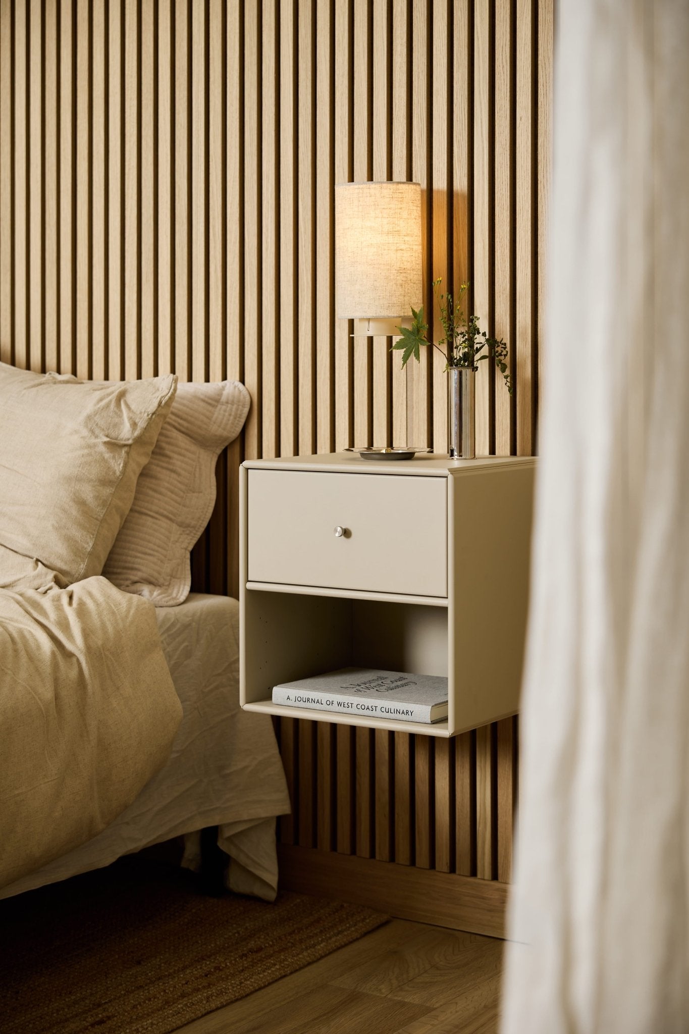 Une commode-bibliothèque WoodUpp Arka de couleur beige, placée près du lit, accueille une lampe, des livres et un vase, et se détache sur un panneau acoustique à lattes de bois WoodUpp Akupanel.