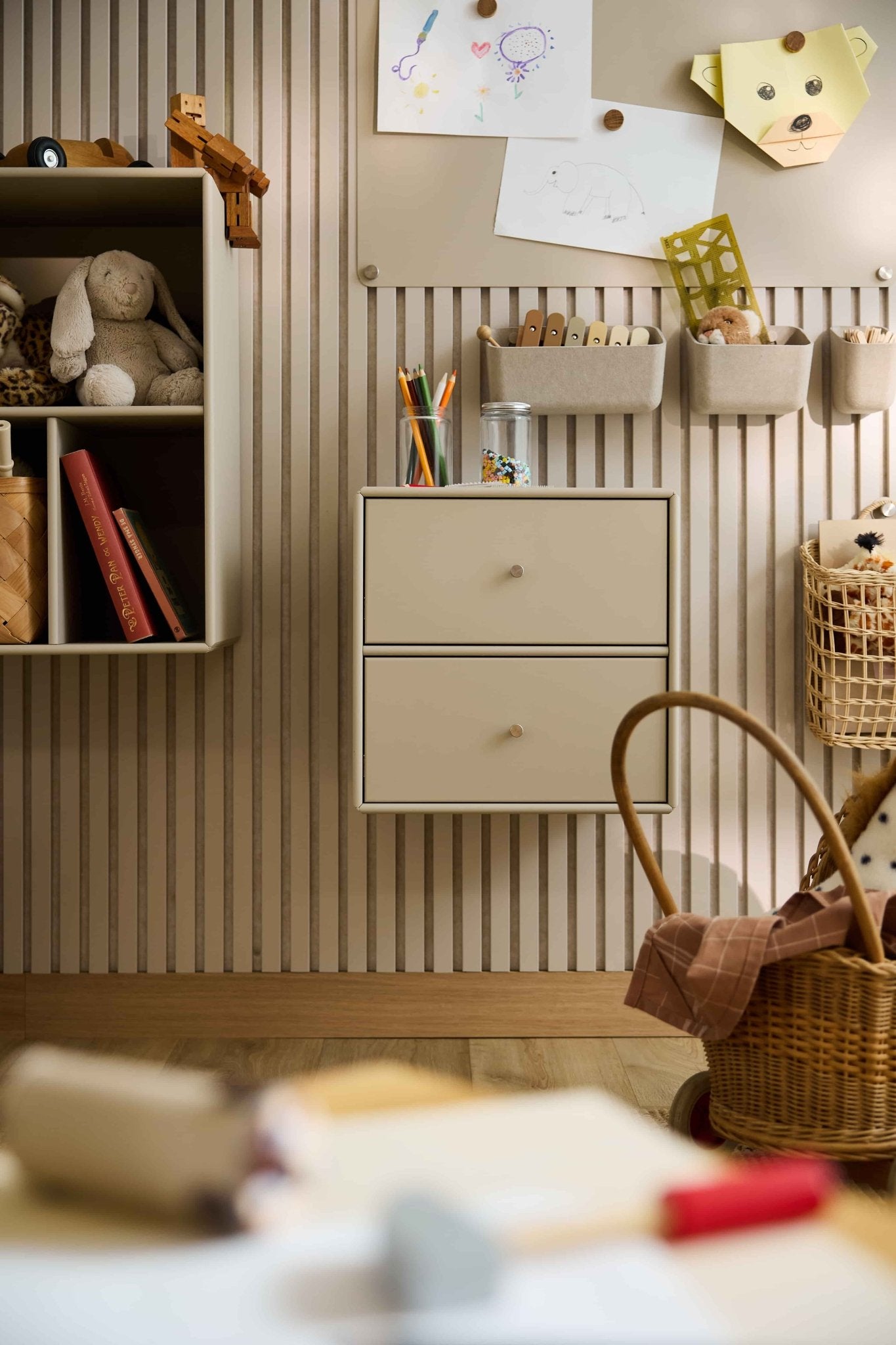 Chambre denfant avec du papier peint à rayures, des jouets, des livres, des paniers et une bibliothèque-commode WoodUpp Arka de couleur beige offrant des rangements fermés.
