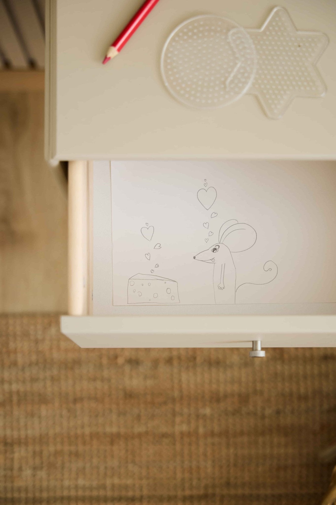Une souris et un morceau de fromage ornés de cœurs décorent un tiroir de la bibliothèque WoodUpp Arka de couleur beige ; un crayon rouge et des panneaux perforés reposent sur le plateau assorti au-dessus.