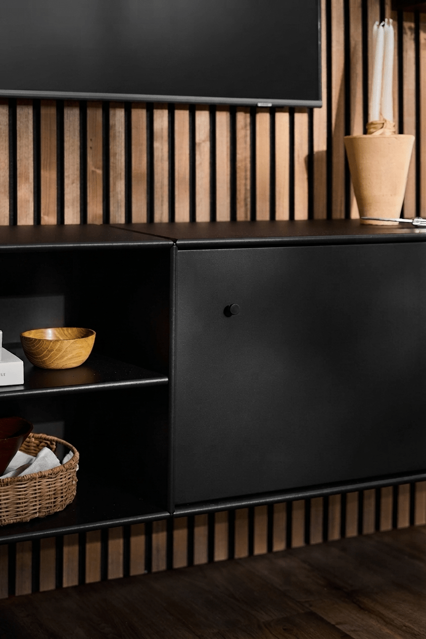 Meuble TV « Black WoodUpp Arka » avec des étagères ouvertes pour les livres et les paniers, un vase beige, un système astucieux de rangement des câbles et un panneau acoustique vertical en bois « Akupanel » en arrière-plan.