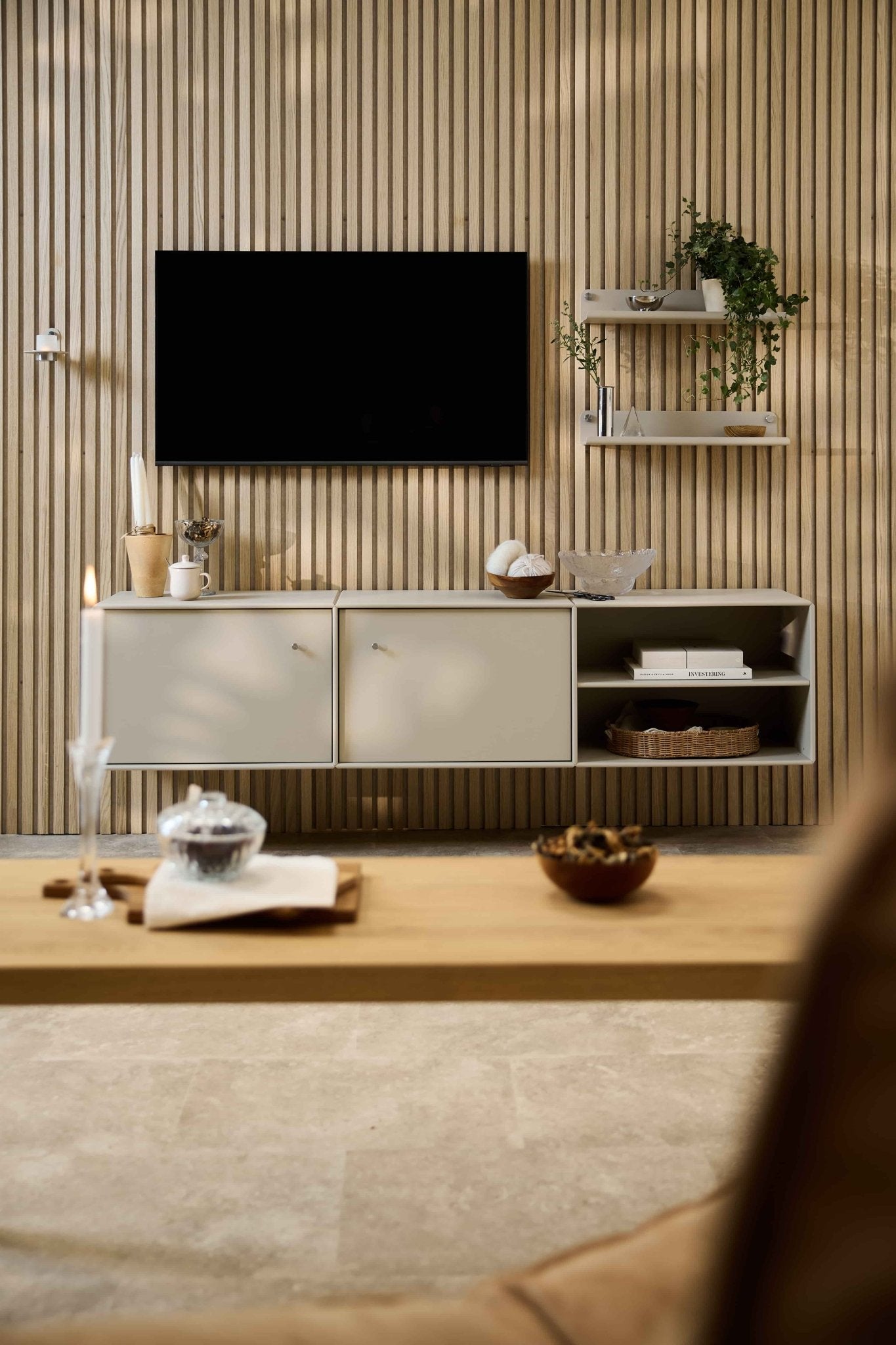 Salon moderne avec le meuble TV « Arka » de WoodUpp, un ensemble de sièges modulables, des étagères de style nordique agrémentées de plantes et dobjets décoratifs, ainsi quune bougie posée sur la table basse.