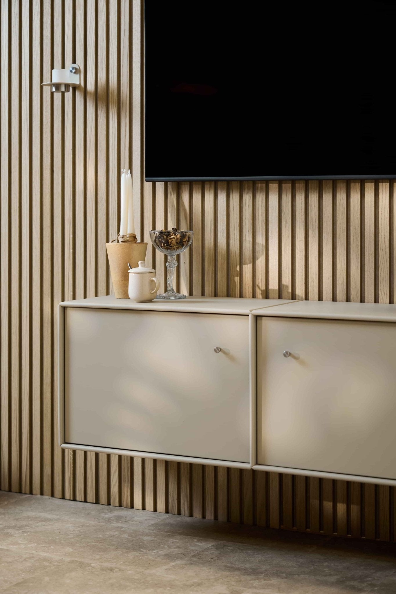 Salon minimaliste avec un meuble TV WoodUpp Arka de couleur beige, un mur recouvert de panneaux acoustiques en bois Akupanel, une télévision fixée au mur, des bougies, un bol en verre, un bocal blanc et des câbles bien rangés.
