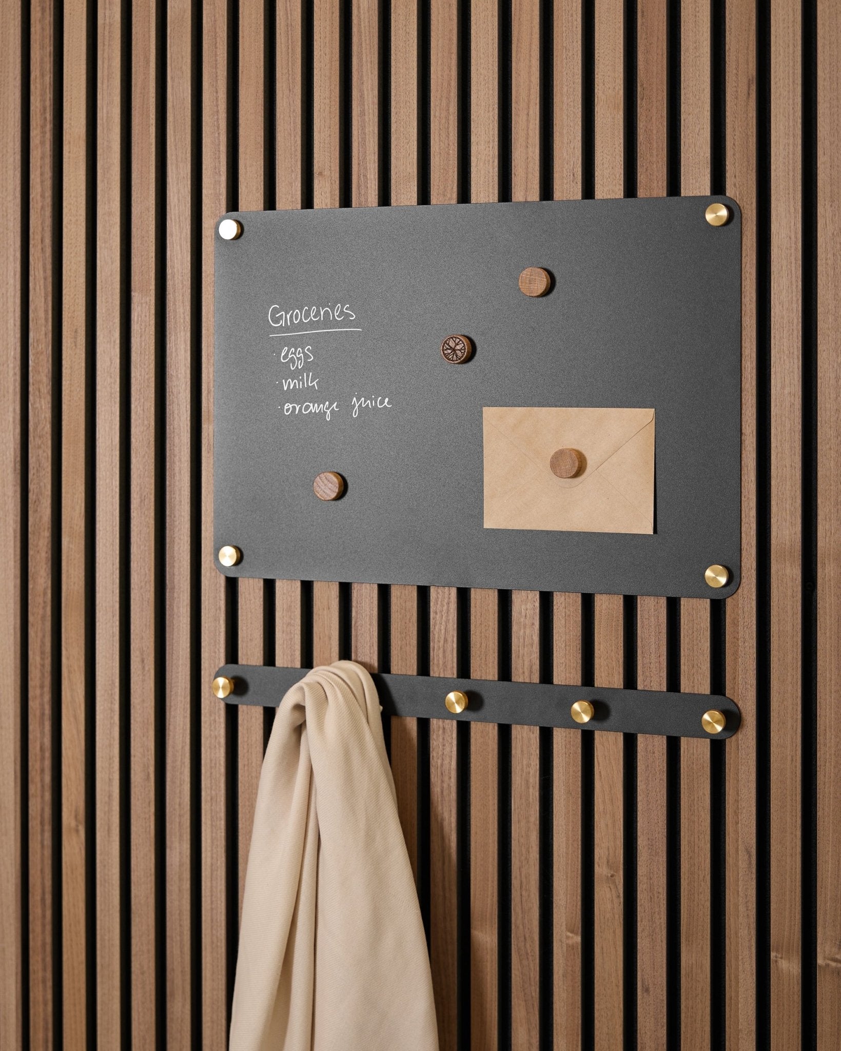 Un tableau magnétique WoodUpp Norr black small accueille une liste de courses, une enveloppe, des aimants et un chiffon beige sur des crochets, monté sur un Akupanel Panneau acoustique.