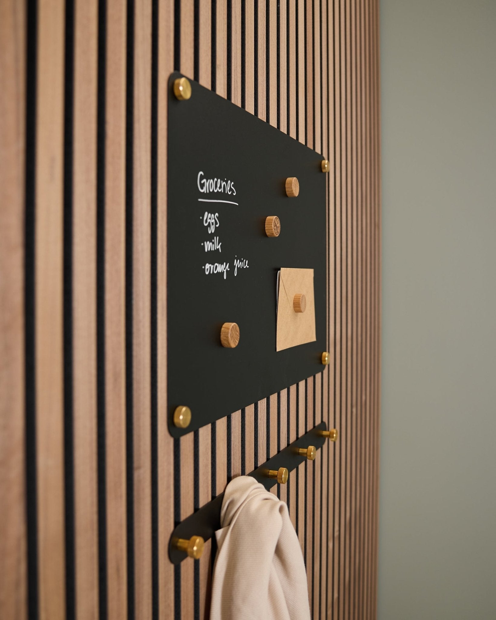 Un tableau magnétique WoodUpp Norr black small, monté sur un panneau acoustique en bois, présente une liste de courses et une note, avec des crochets en dessous qui supportent un tissu léger.