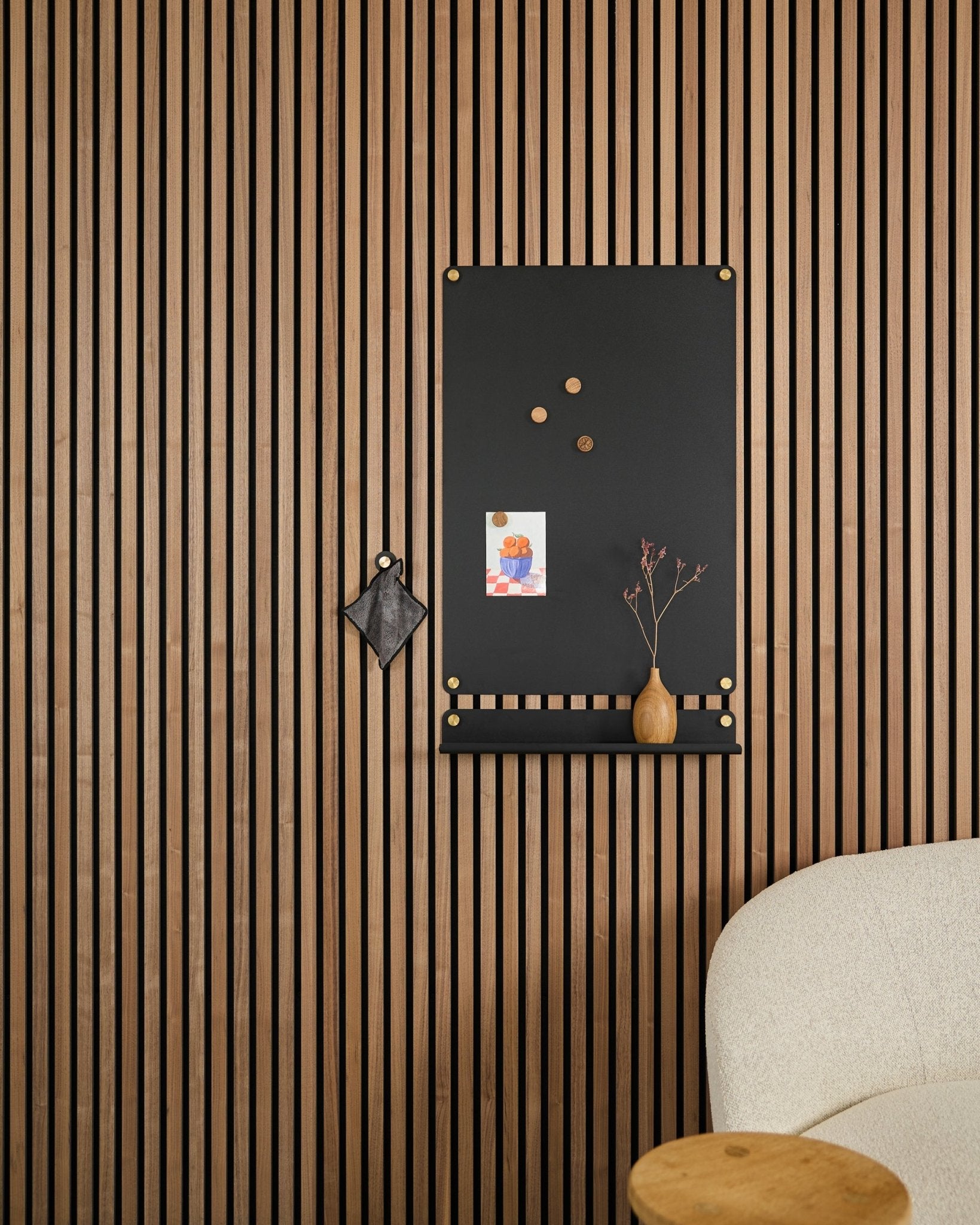 Un large black Norr tableau magnétique de WoodUpp, conçu pour les panneaux acoustiques, est suspendu à côté d’une chaise blanche rembourrée, avec des notes, un vase et un petit objet décoratif.