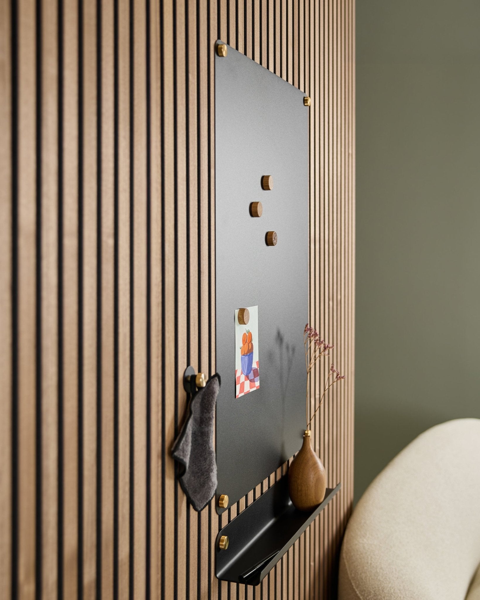 Le Norr large black tableau magnétique acoustique WoodUpp avec des aimants en bois, un tableau d’affichage, une étagère et un vase est fixé au mur à côté d’une chaise beige.