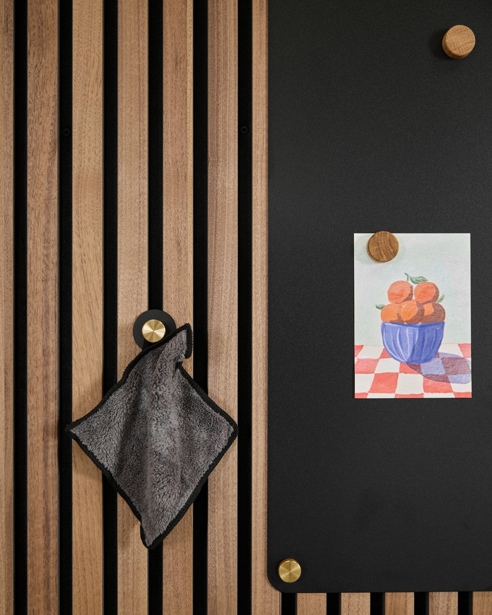 Une serviette small est suspendue à un panneau acoustique à lattes de style nordique, à côté d’un tableau black présentant un dessin d’oranges dans un bol, fixé avec deux aimants dorés.