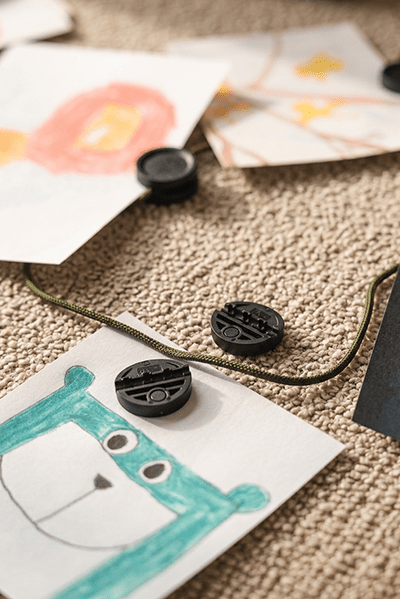 Des dessins d’enfants sont suspendus au Vindar | Gallery String de WoodUpp avec des clips en néodyme ; une illustration verte d’ours repose sur la moquette beige sous le Panneau acoustique.