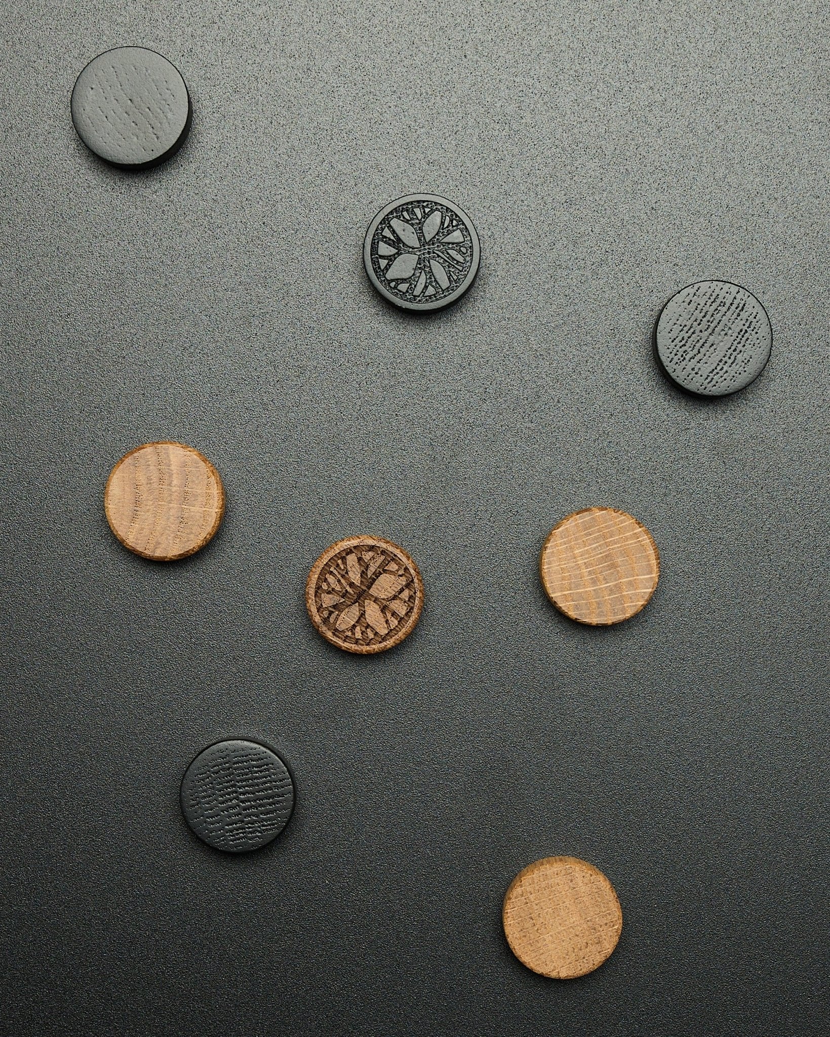 Quatre aimants ronds black oak du lot d’aimants WoodUpp Norr, disposés librement sur une surface sombre texturée, conçus comme accessoires Akupanel.