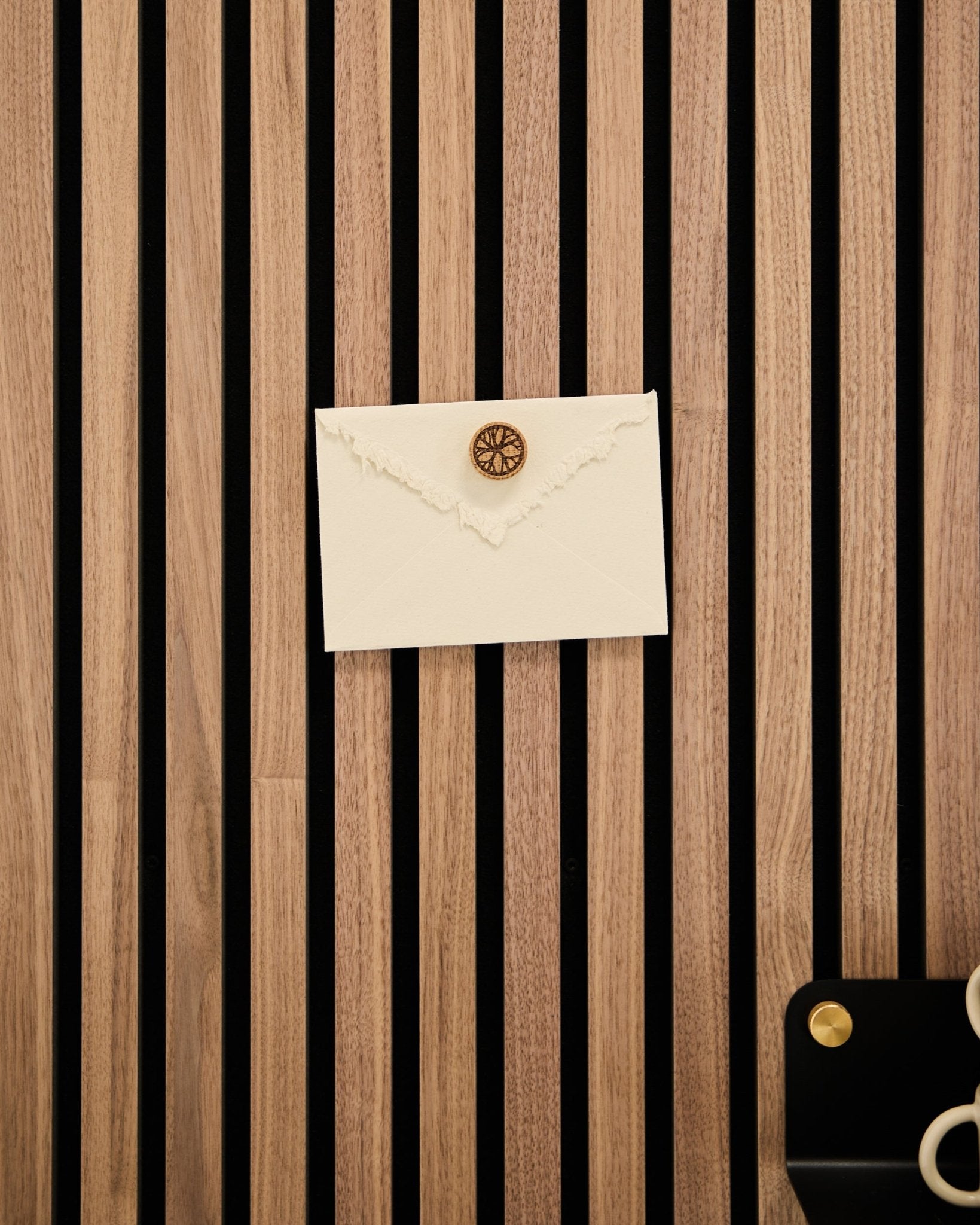 Une enveloppe crème avec un sceau de cire est fixée sur un panneau acoustique à lattes en chêne WoodUpp à l’aide du set d’aimants pour posters Norr en Natural Oak ; des ciseaux et un plateau sont posés à côté.