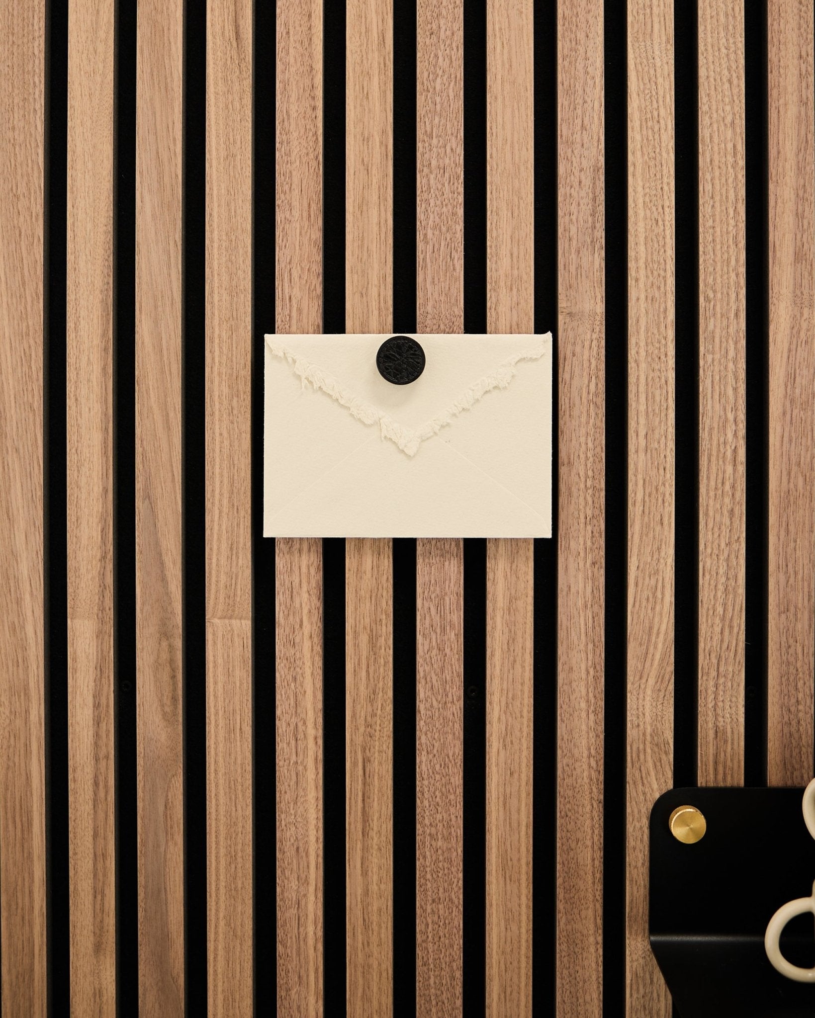 Une enveloppe crème est fixée sur un panneau acoustique WoodUpp à lattes de chêne Akupanel Panneau acoustique à l’aide du set de Poster Magnet Norr en Black Oak ; des ciseaux reposent en bas à droite.
