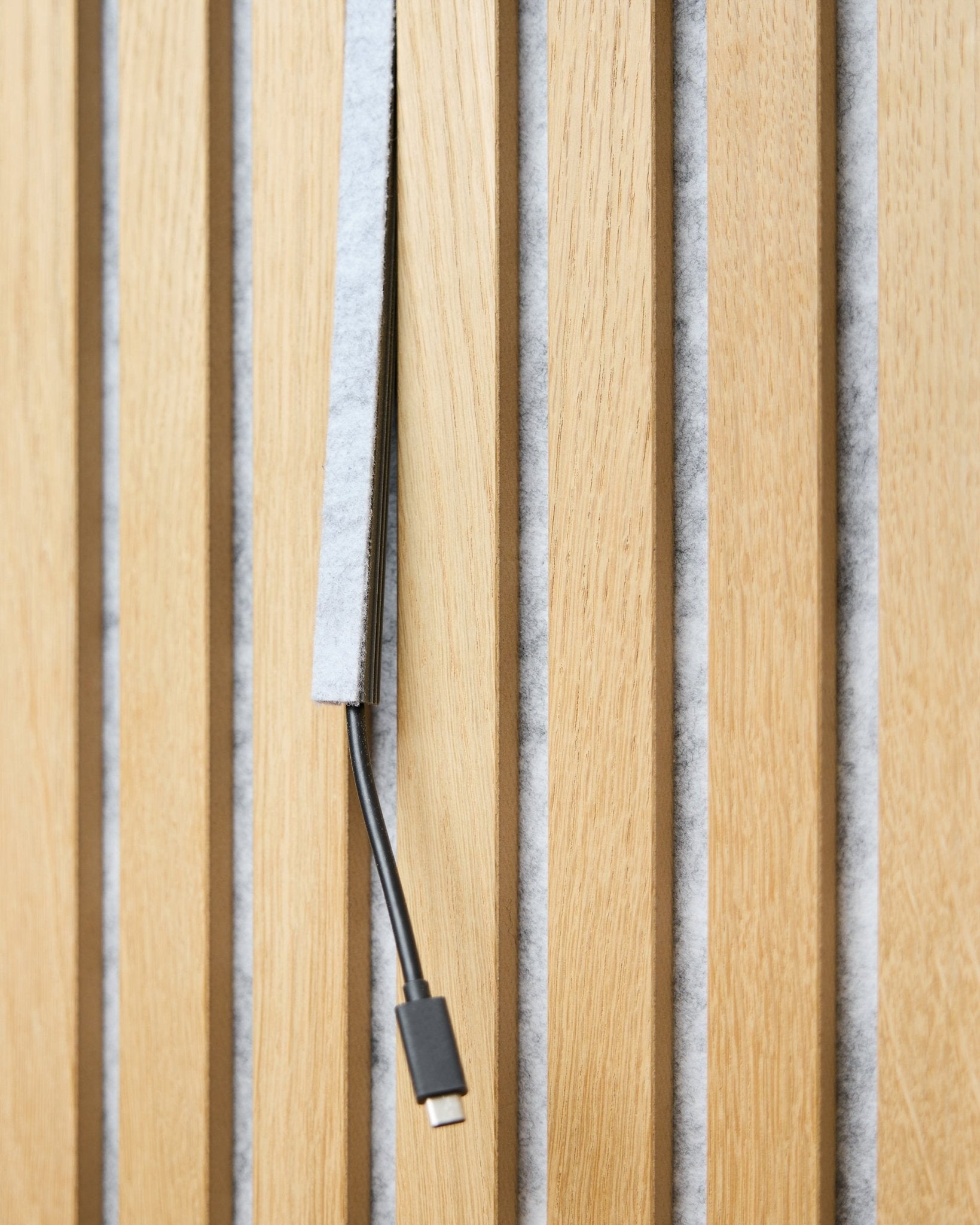 Un câble USB-C black passe dans un cache-câble Pop-on WoodUpp Dupps grey, entre des lattes acoustiques en bois vertical avec support en feutre grey.