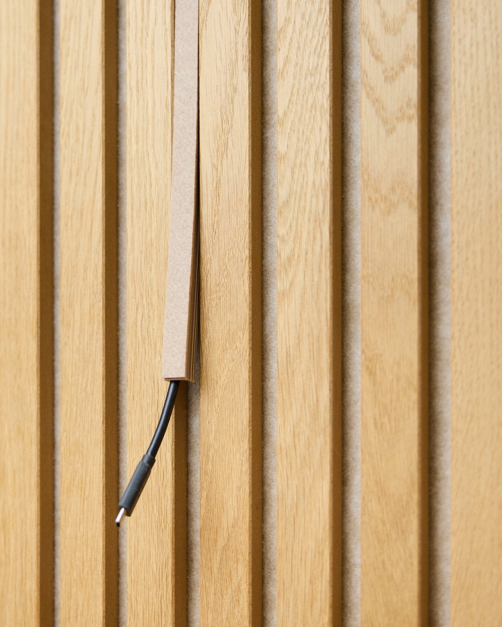 Un câble black avec gestion des câbles est partiellement dissimulé dans un cache-câble Pop-on WoodUpp Dupps Beige, monté sur des lattes acoustiques en bois vertical Akupanel.