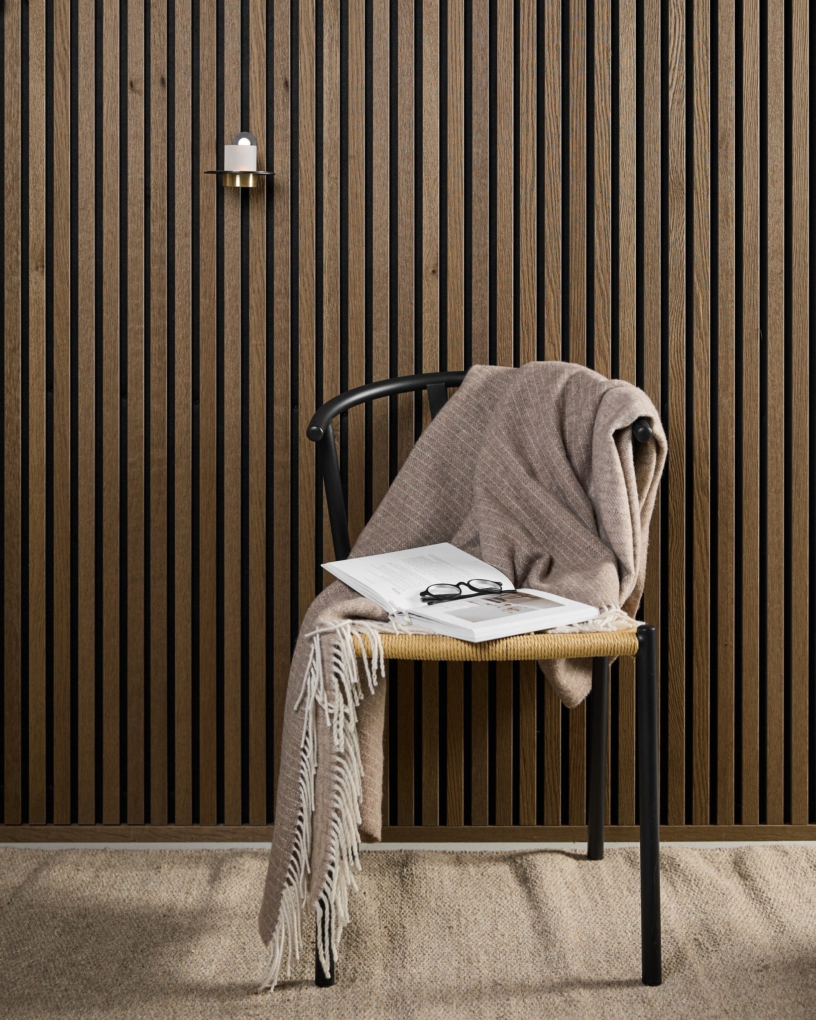 Une chaise moderne avec un plaid beige, un livre ouvert et des lunettes est placée près d’un panneau acoustique en bois Akupanel et d’un bougeoir WoodUpp Norr black et brass.