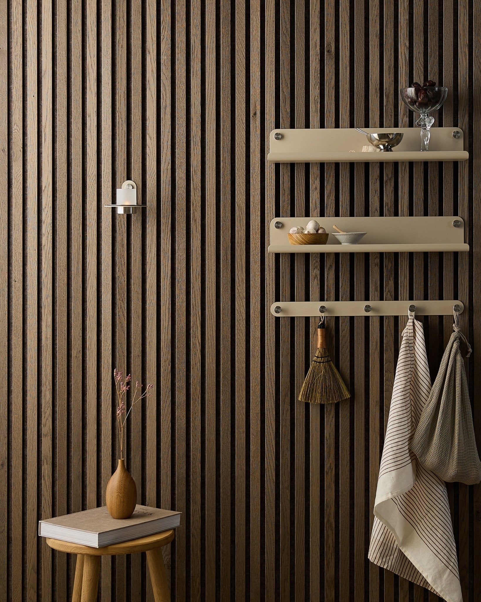 Un tabouret en bois avec des fleurs séchées se trouve à côté d’un Akupanel Panneau acoustique WoodUpp, près d’étagères beige accueillant de la vaisselle, une brosse, des serviettes et un bougeoir Norr.