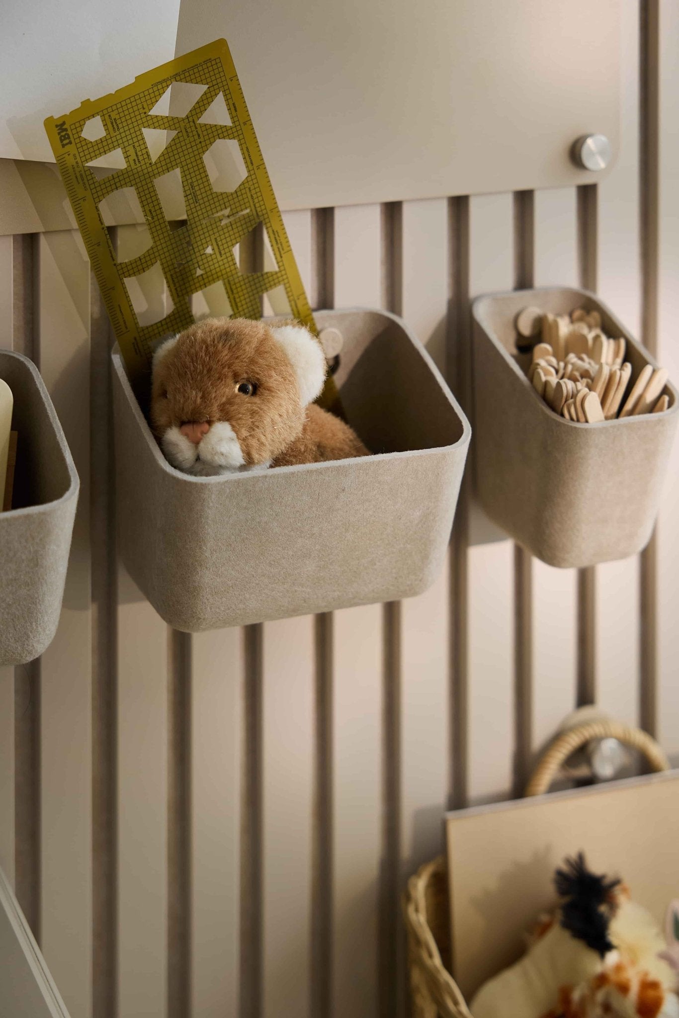 Les bacs en feutre « WoodUpp Luva » de couleur beige contiennent des jouets, une règle jaune et des bâtonnets en bois, permettant un rangement organisé sur un panneau mural acoustique à lattes rayé.