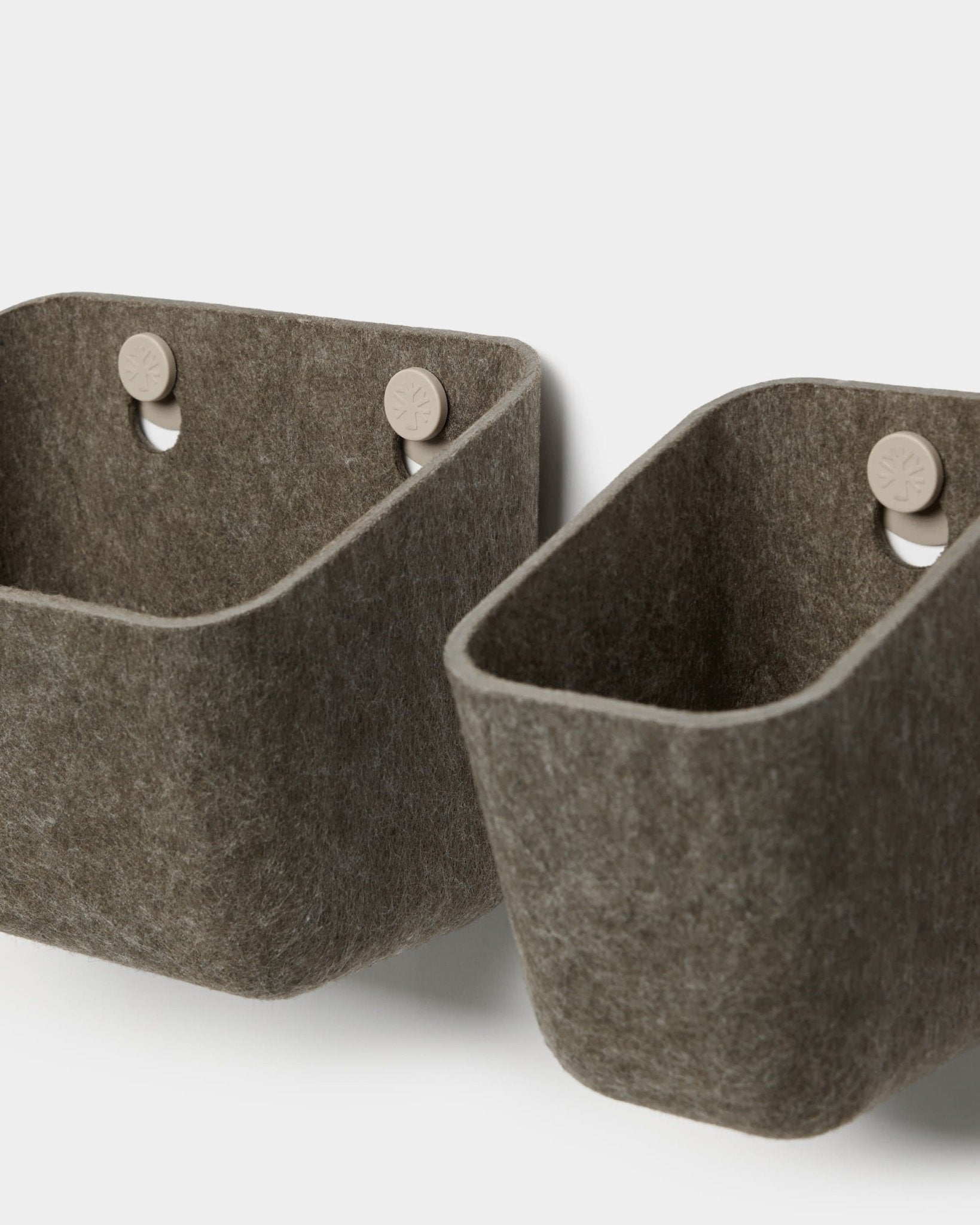 Deux rangements en feutre Luva, de couleur gris foncé chaud, signés WoodUpp, sont suspendus à des crochets beiges fixés à un panneau acoustique, offrant ainsi un rangement mural minimaliste aux angles arrondis.