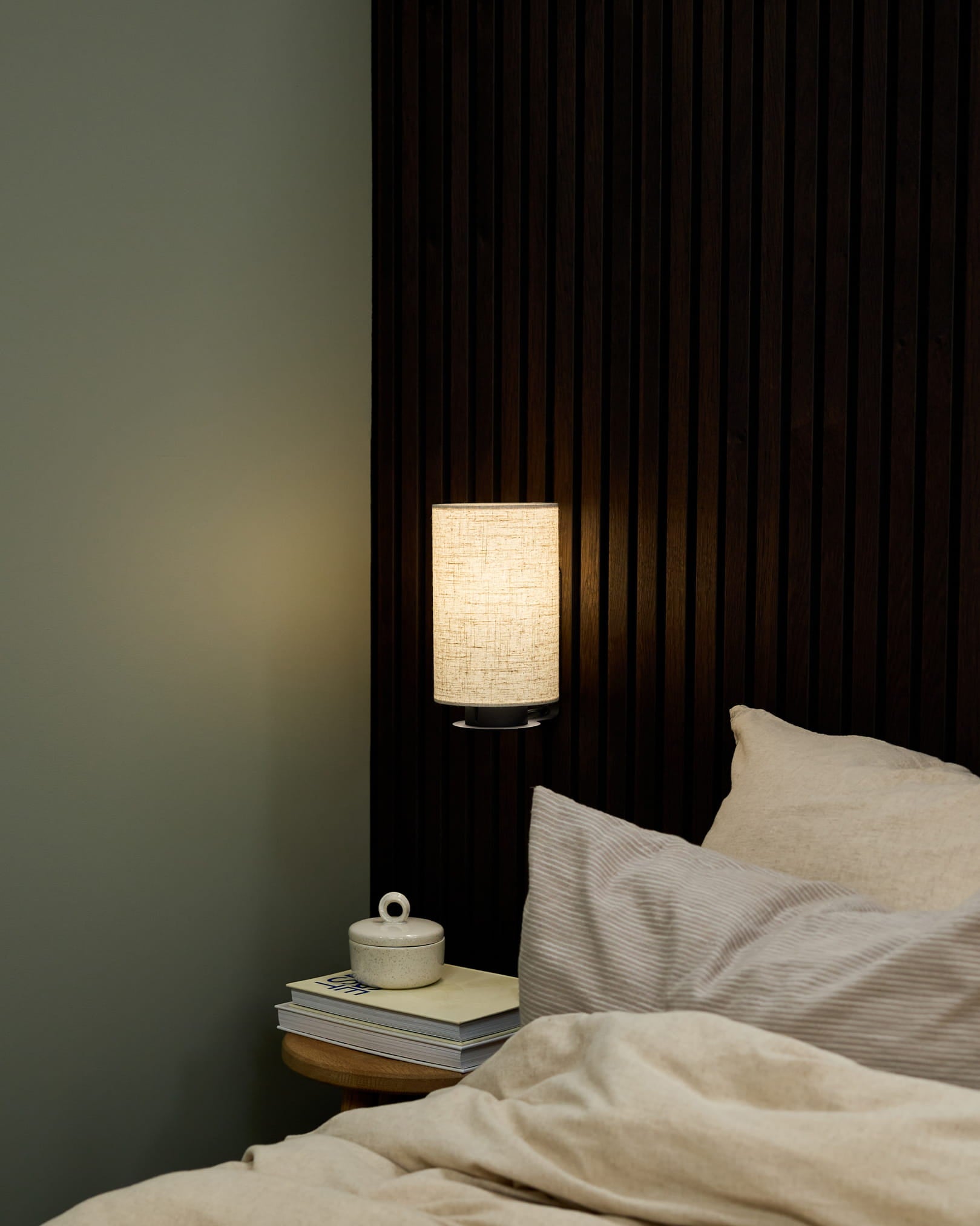 Coin chambre avec une applique murale Feya de WoodUpp en Black/Beige, du linge de lit beige et une table de chevet avec des livres et une tasse en céramique avec couvercle.