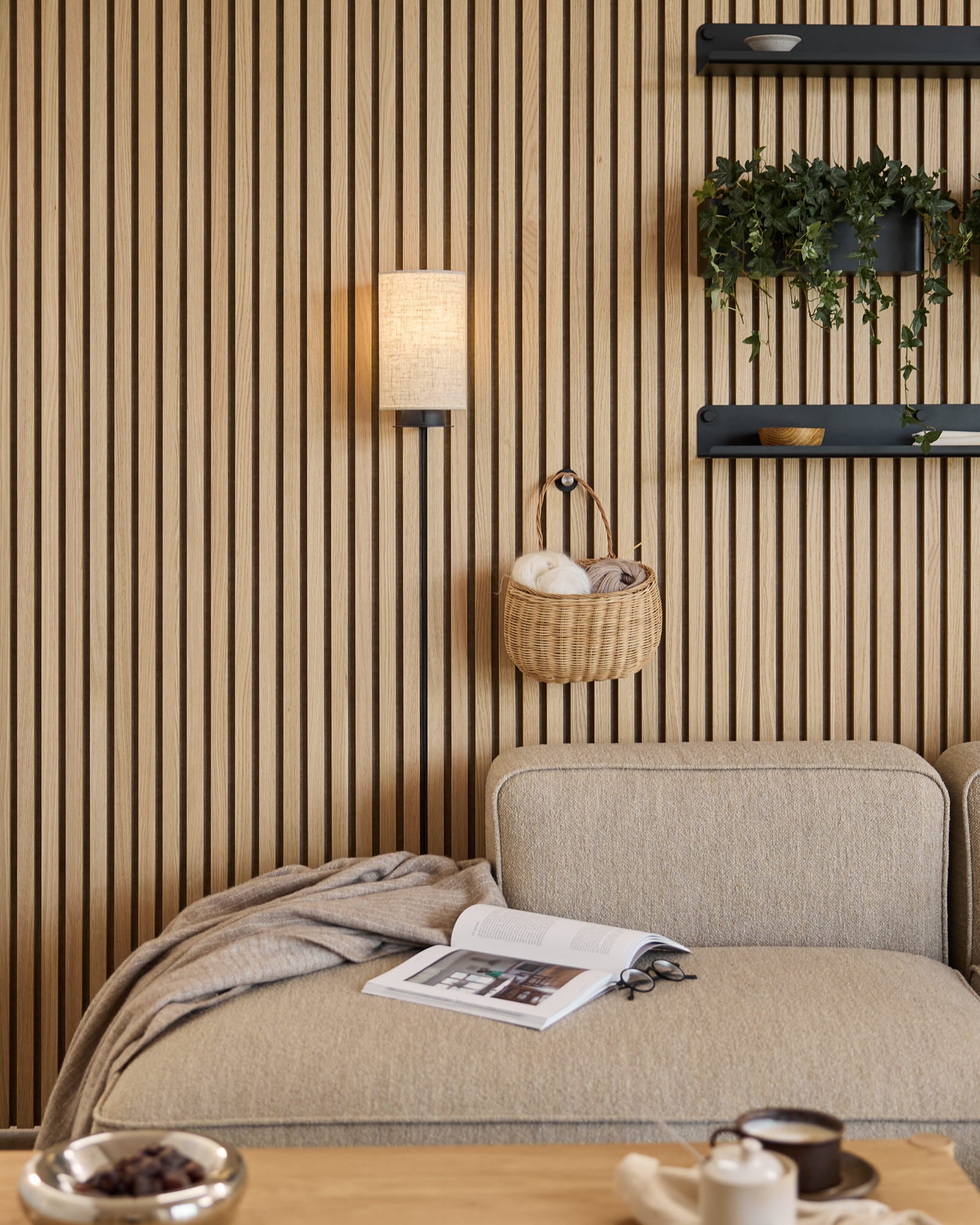 Canapé Beige avec plaid et livre, à côté d’un mur acoustique à lattes WoodUpp Akupanel, d’étagères avec plantes et objets déco, et d’une applique murale Feya en Black/Beige.