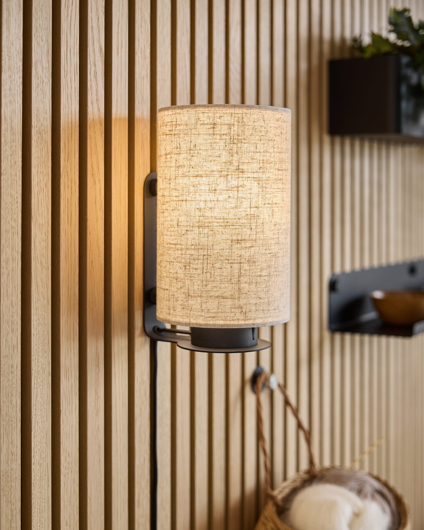 Une applique murale moderne avec un abat-jour en tissu projette une lumière chaude sur des panneaux de bois acoustique Akupanel texturés, complétés par des étagères minimalistes et un panier tressé.