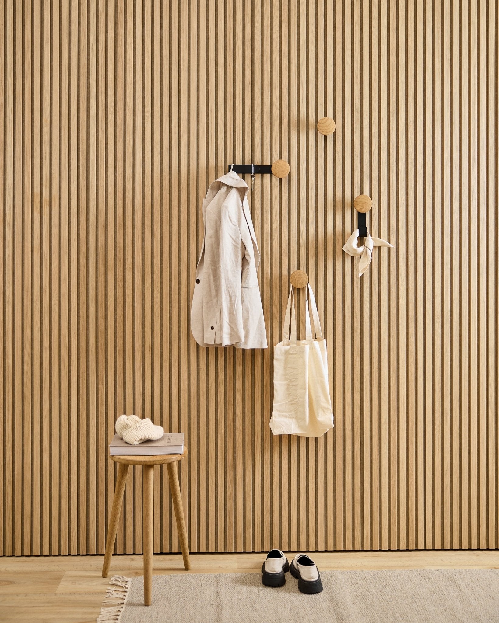 Entrée minimaliste avec le porte-manteau WoodUpp Linn Vertical en natural oak, accompagné d'un manteau beige, d'un cabas, d'une écharpe, d'un tabouret avec des chaussons et de chaussures black et blanches.
