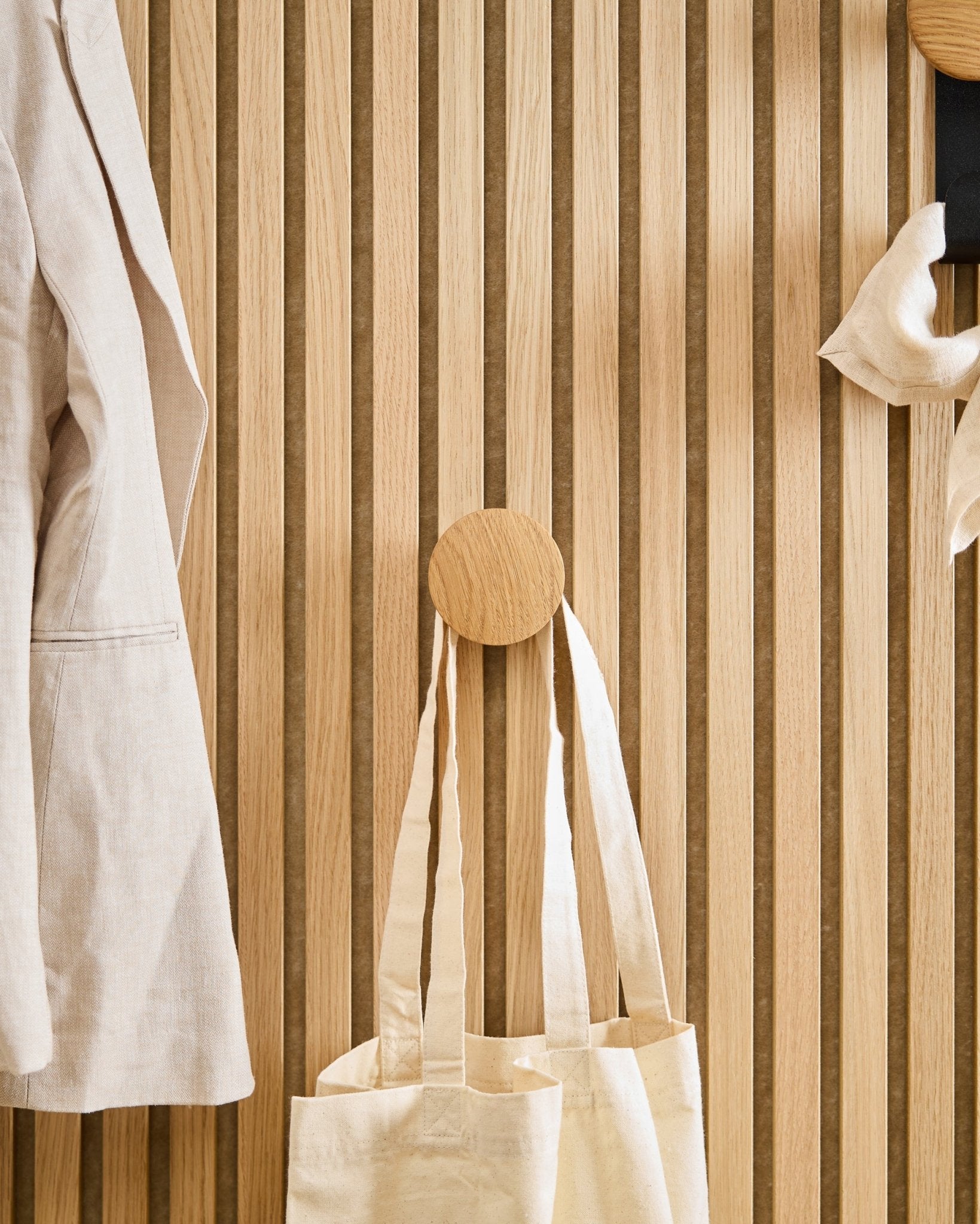 Un manteau beige et un cabas sont suspendus au Linn I Single Hanger en Natural Oak de WoodUpp, monté sur un panneau acoustique à lattes de bois vertical Akupanel.