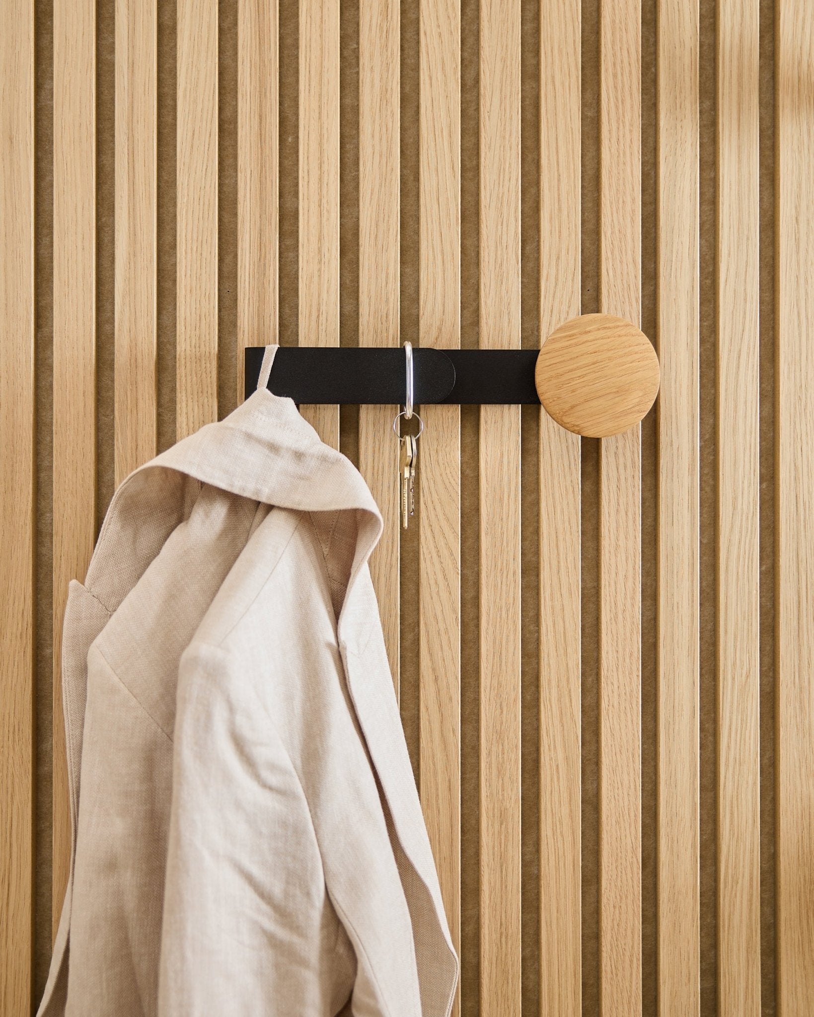 Un blazer beige et un trousseau de clés sont suspendus au Linn Horizontal Hanger de WoodUpp en natural oak, installé sur un panneau acoustique en bois Akupanel natural oak.