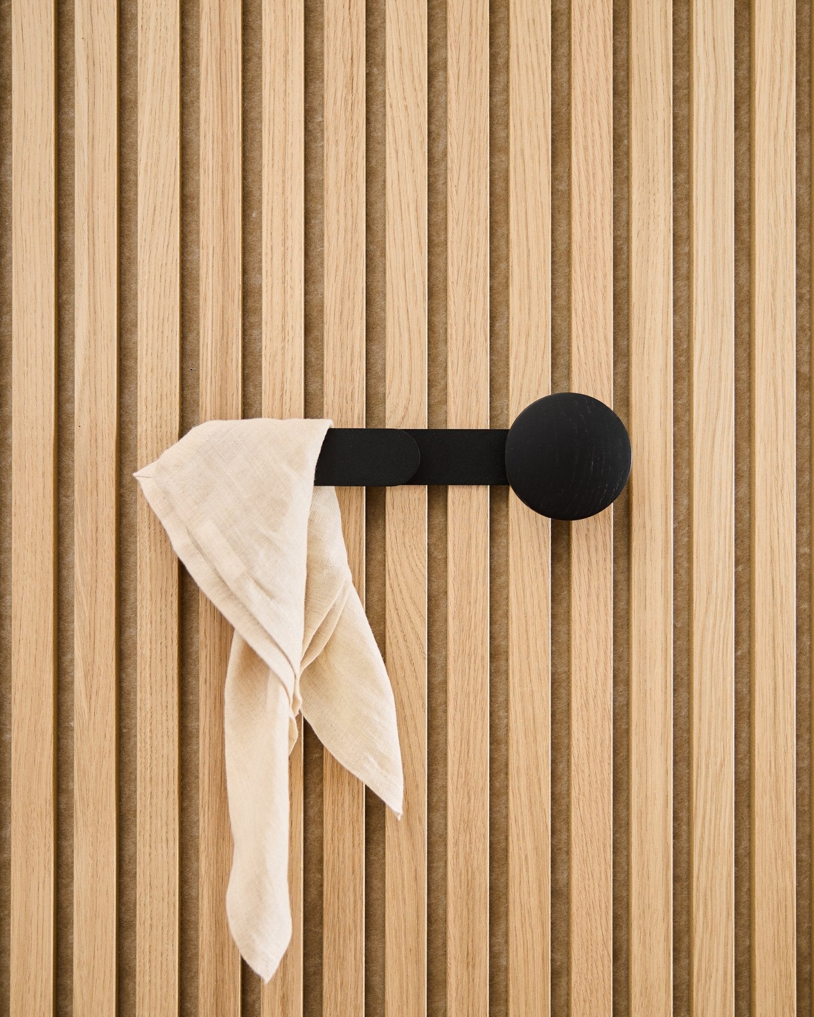 Un tissu beige est drapé sur le porte-manteau WoodUpp Linn Horizontal en Black Oak, fixé à un panneau acoustique à lattes vertical.
