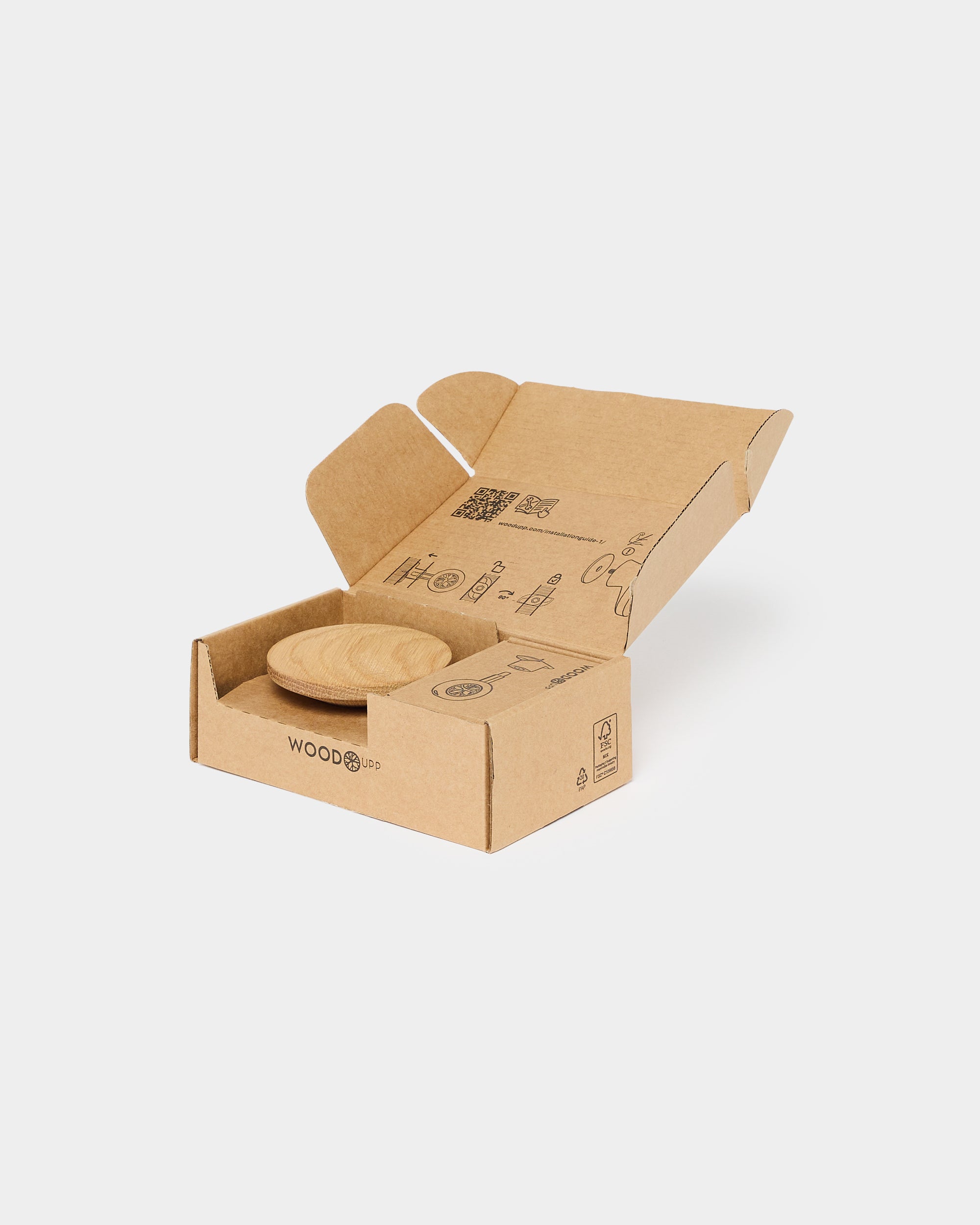 A small round wooden token sits in an open brown cardboard box with printed symbols and text on the inner flaps, against a white background.