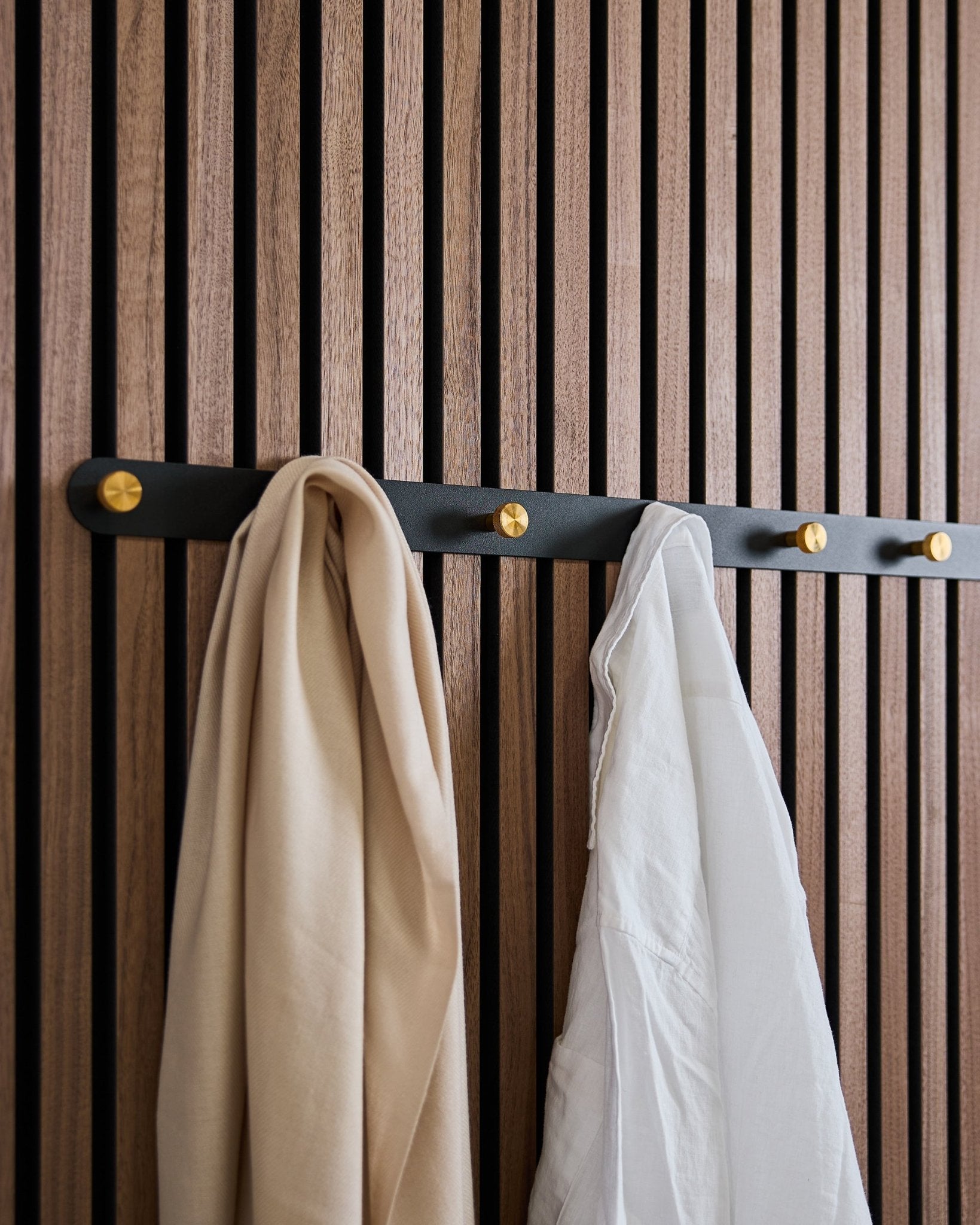 Le porte-manteau mural WoodUpp Norr en black/brass (X7), avec une écharpe beige et une chemise blanche, est suspendu sur un panneau acoustique mural en bois à lattes au style nordique.