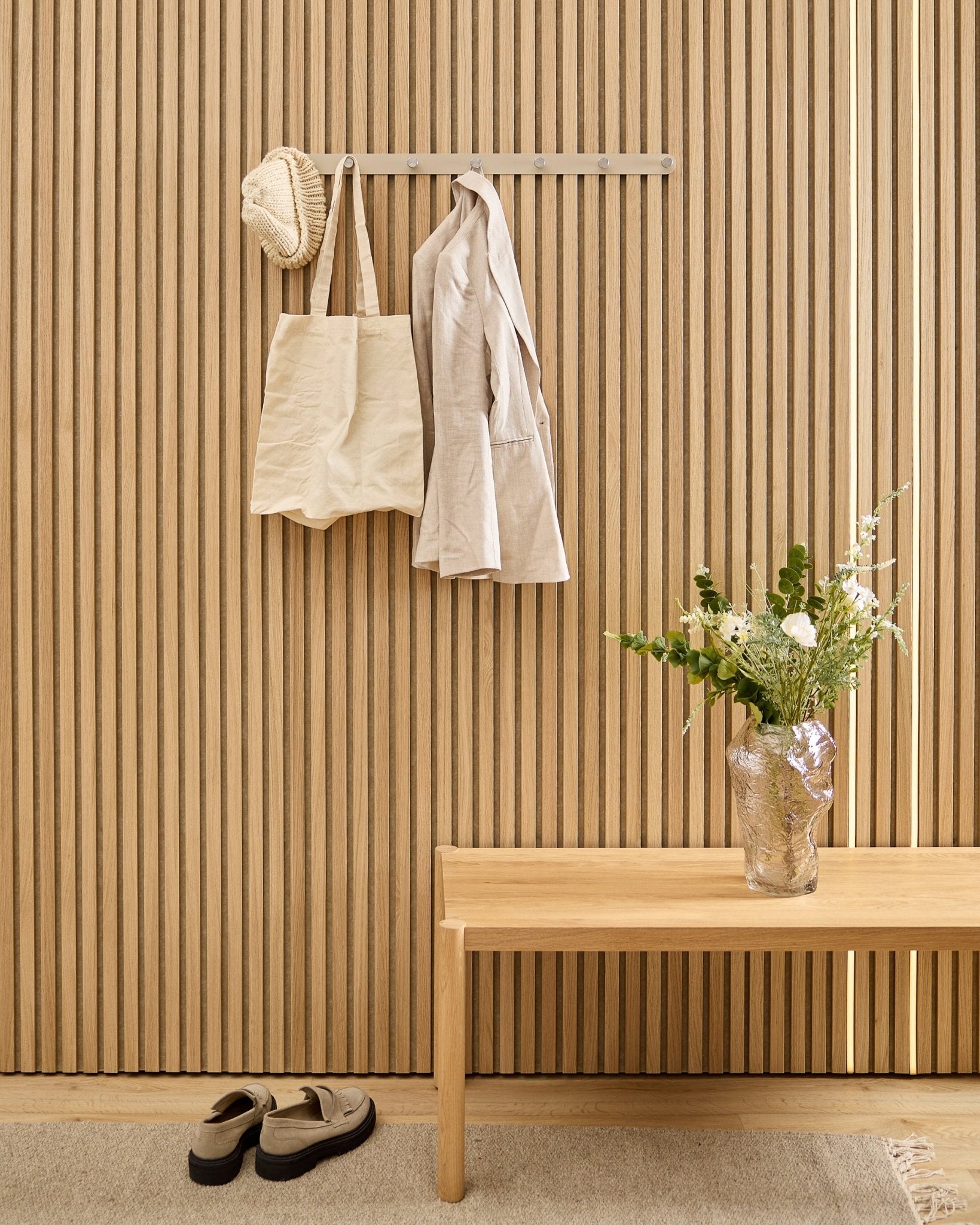 Un sac beige, un chapeau et une chemise sont suspendus à la patère murale WoodUpp Norr au-dessus de chaussures, avec un banc et un vase à côté du panneau acoustique à lattes rayées.