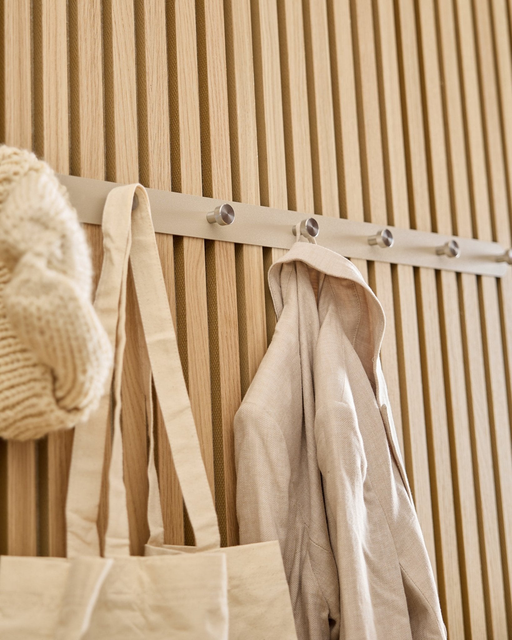 Un cabas beige, un bonnet en maille et une chemise en lin sont suspendus à la patère murale WoodUpp Norr (Beige/Acier inoxydable Steel, X7) fixée sur un panneau à lattes Akupanel en bois clair.